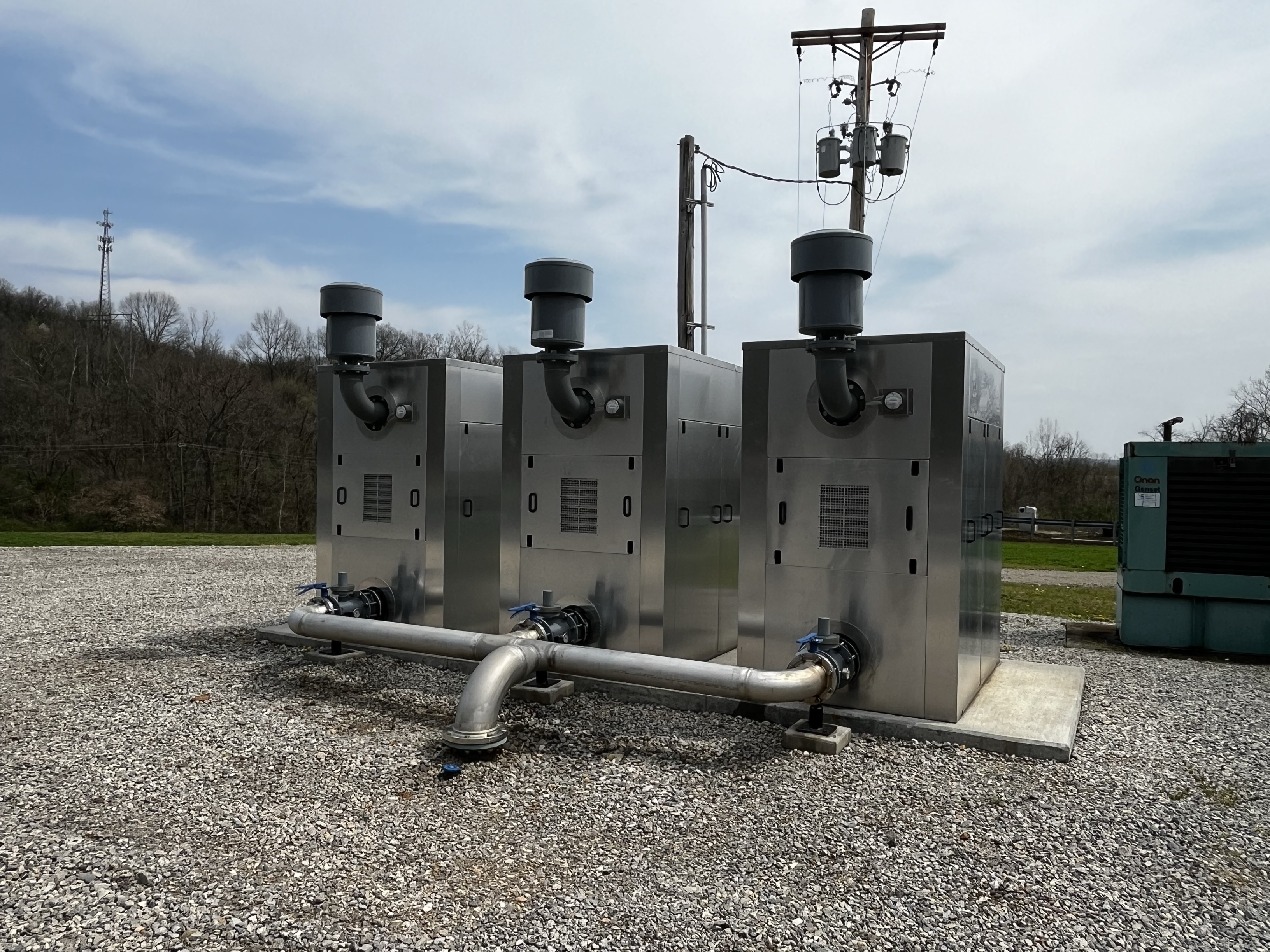 Three stainless steel industrial enclosures with exhaust fans and large pipes on a gravel pad.