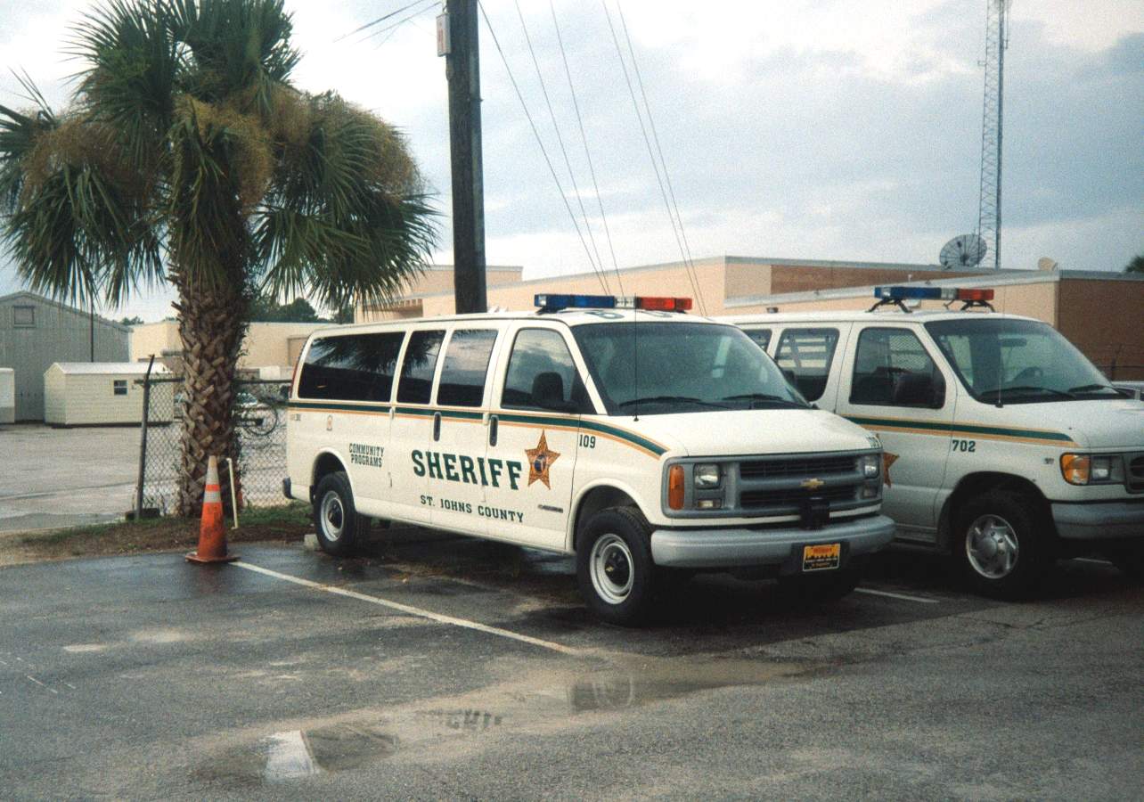 Two white sheriff's office vehicles with emergency light bars parked near a palm tree.