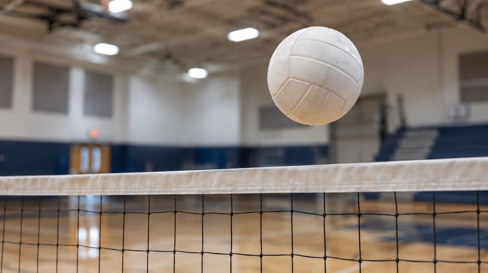 A white volleyball suspended above a net on a hardwood gymnasium court.