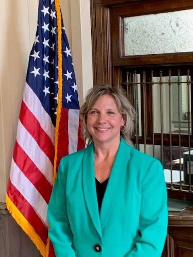 A woman in a teal blazer smiles in front of an American flag in an official building.