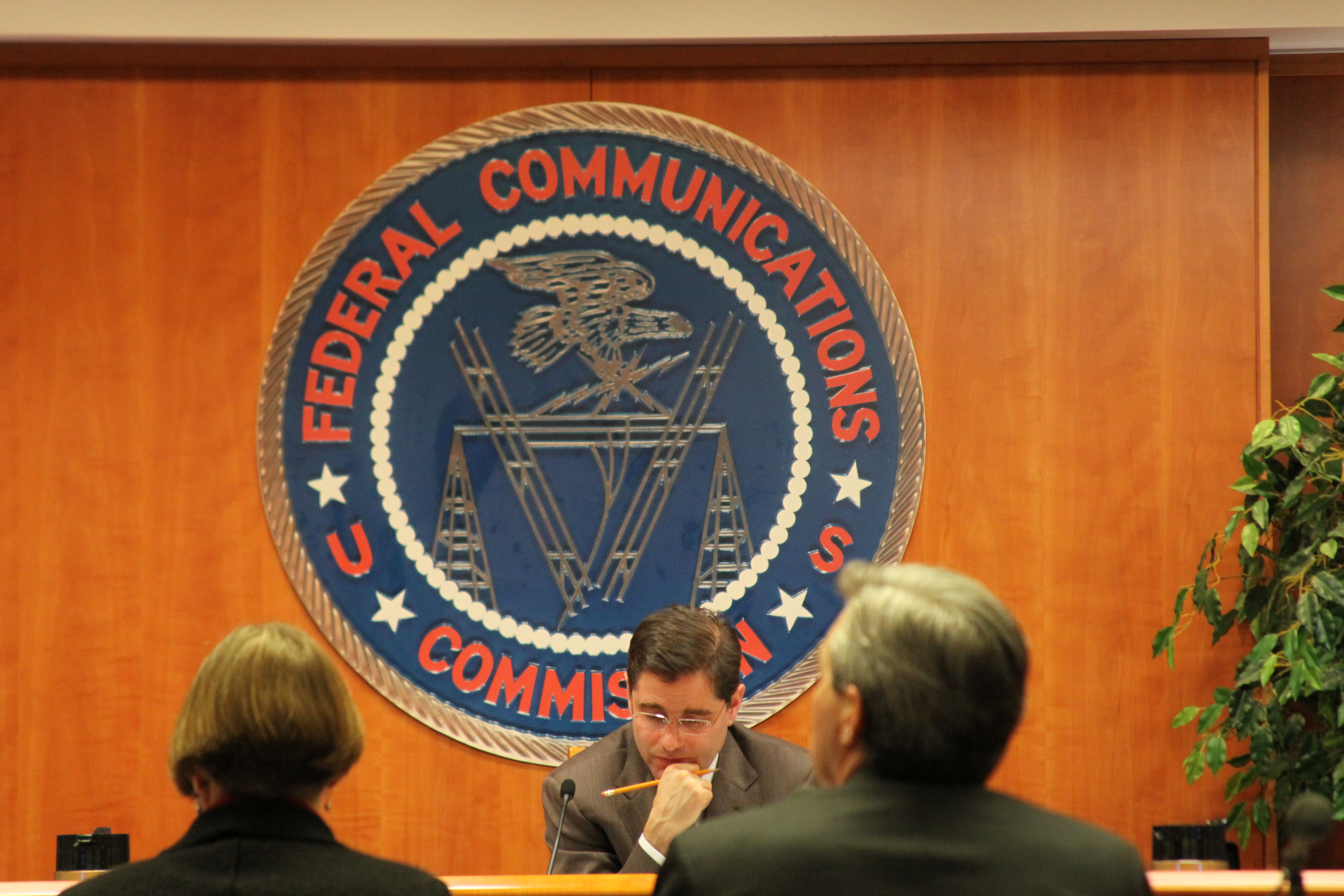 Commissioner seated at a hearing table before a large Federal Communications Commission seal.