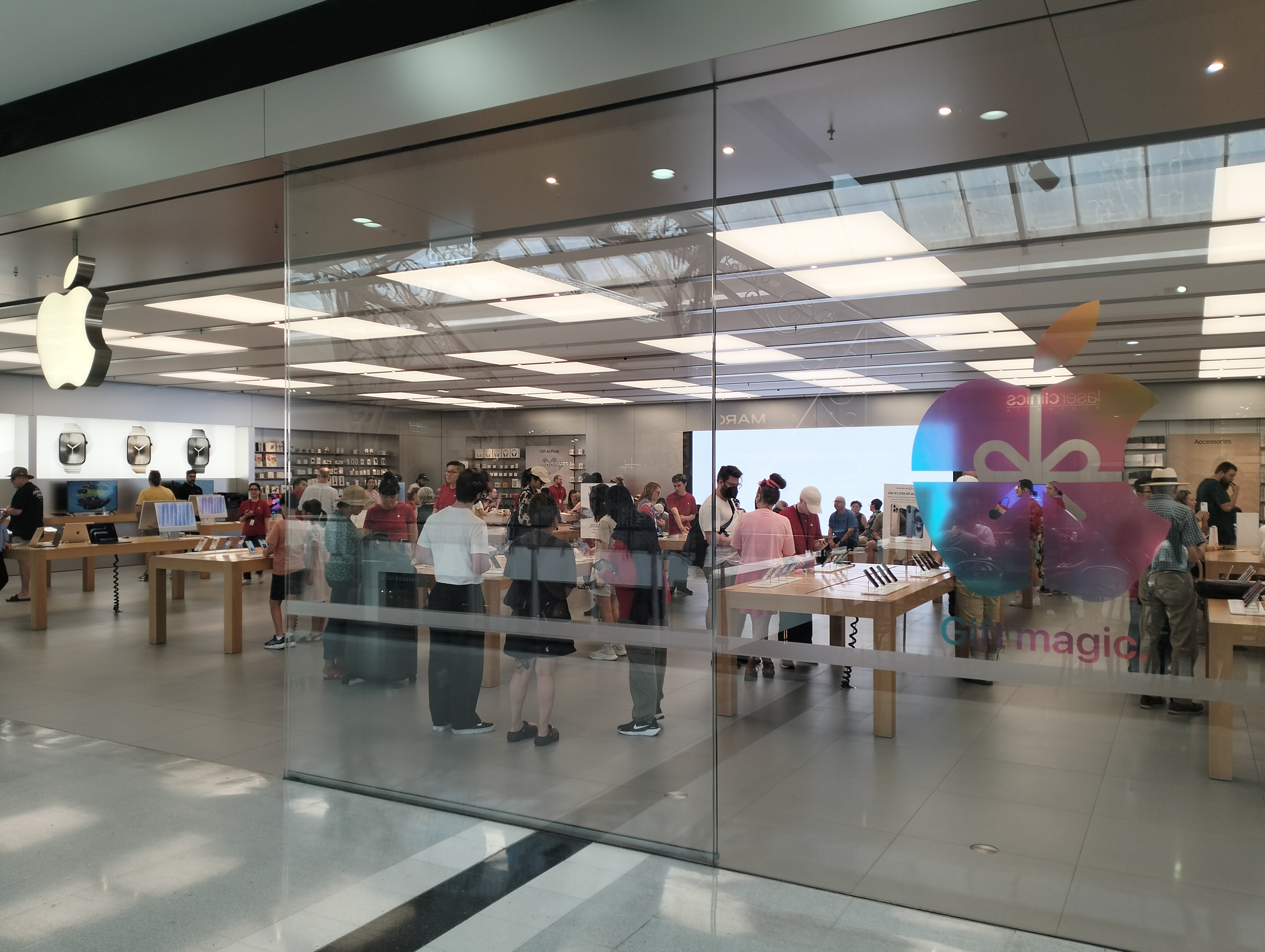 Customers browse Apple products on wooden tables inside a glass-fronted Apple Store.