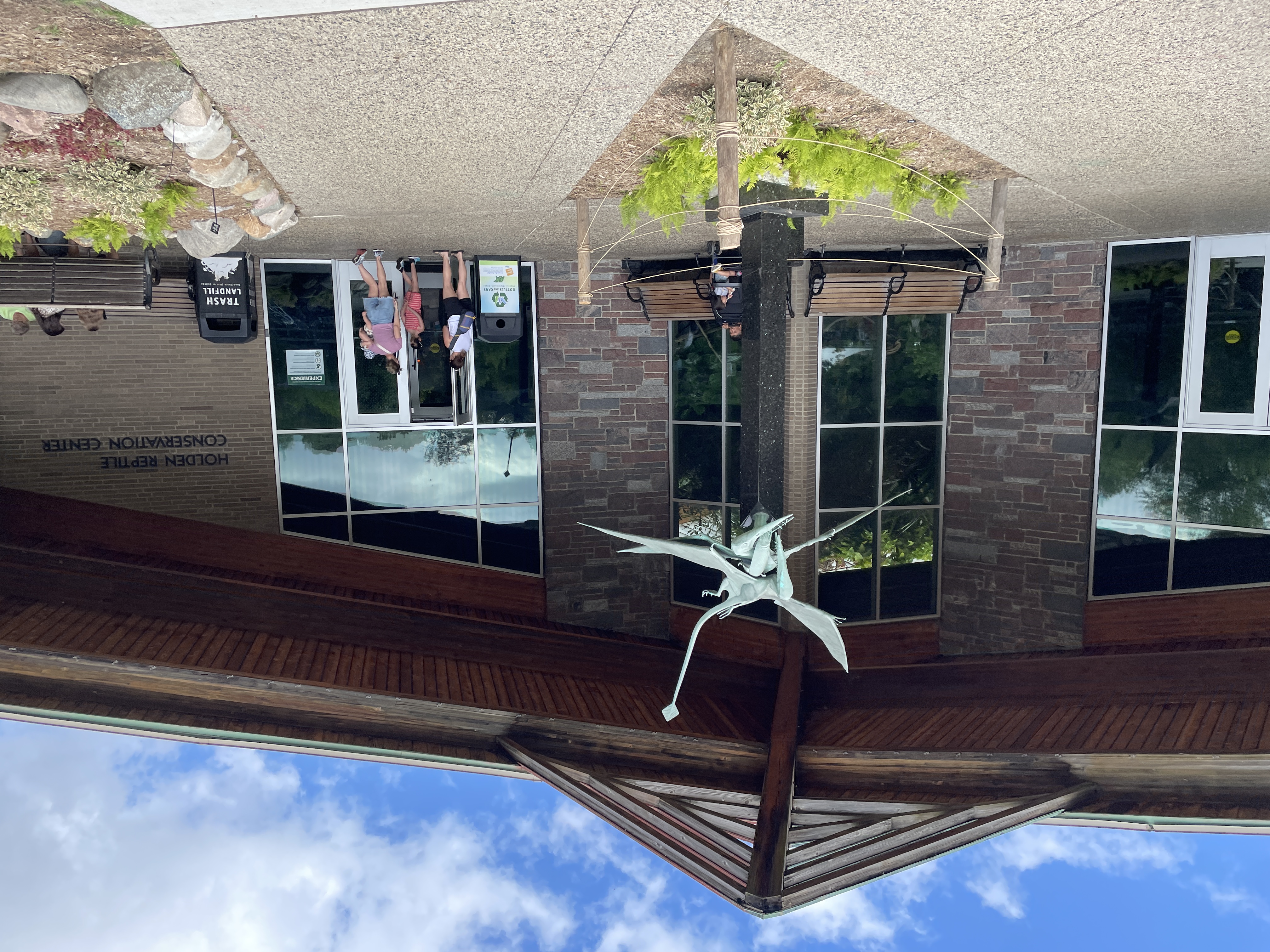 A winged reptile sculpture stands outside a brick building labeled “Holden Reptile Conservation Center,” with visitors near the entrance.