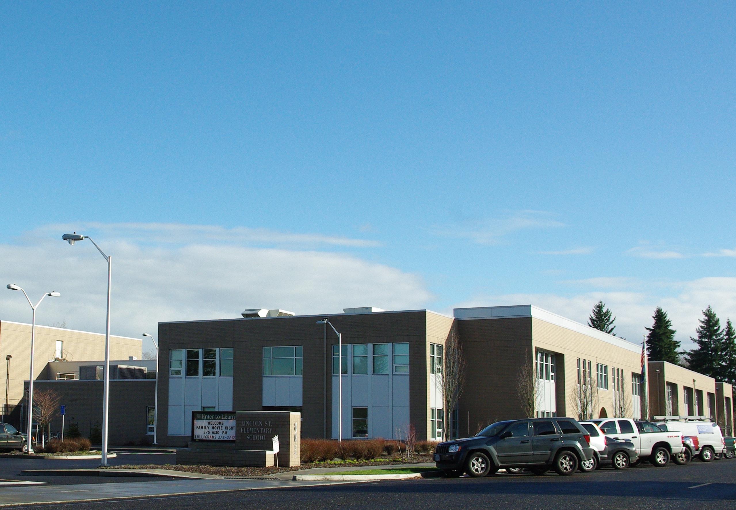 Two-story school building with a parking lot, street lights, and evergreen trees under a blue sky.