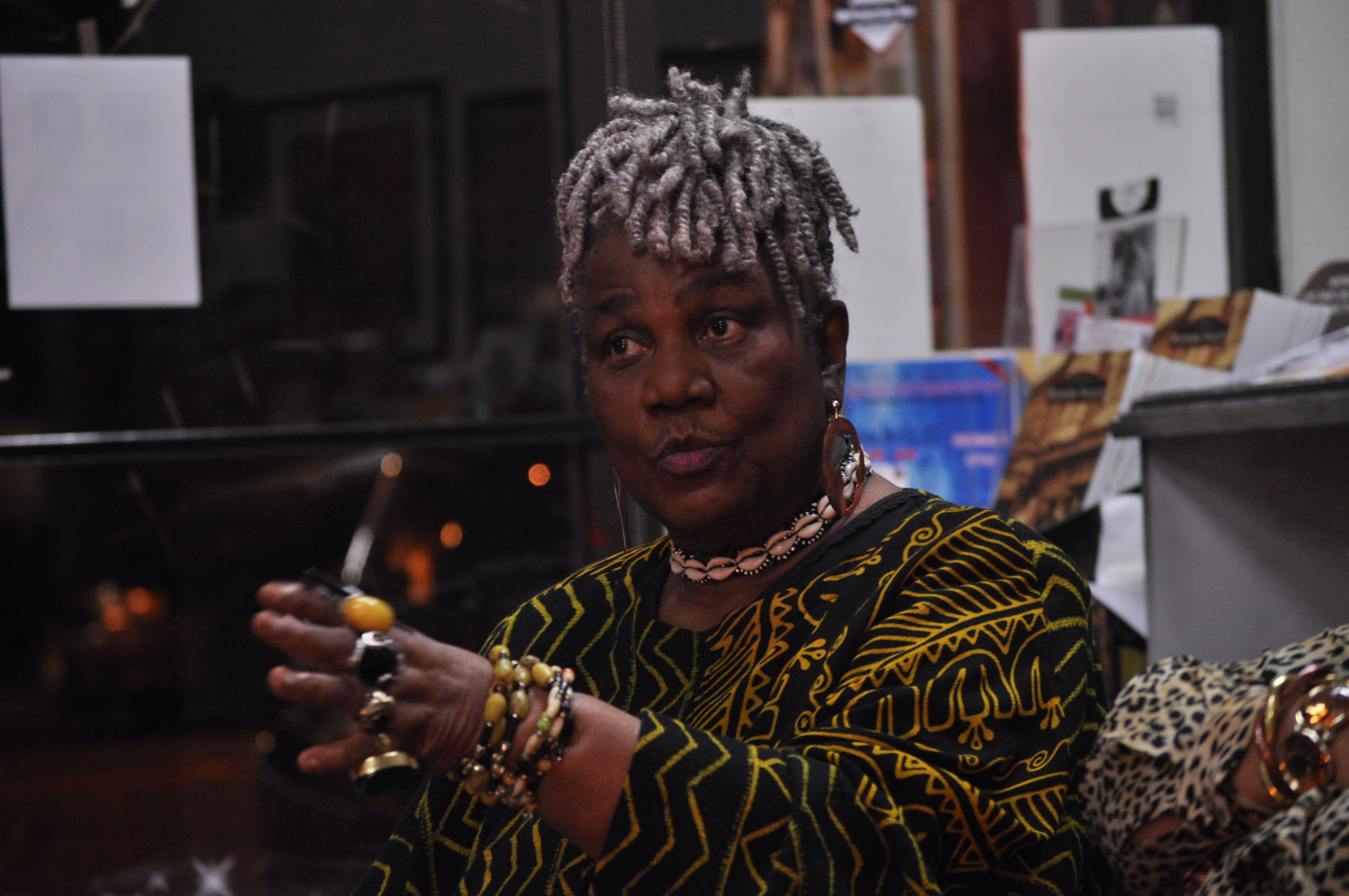 Older woman with gray locs and patterned top seated in a bookstore, hands raised in conversation.