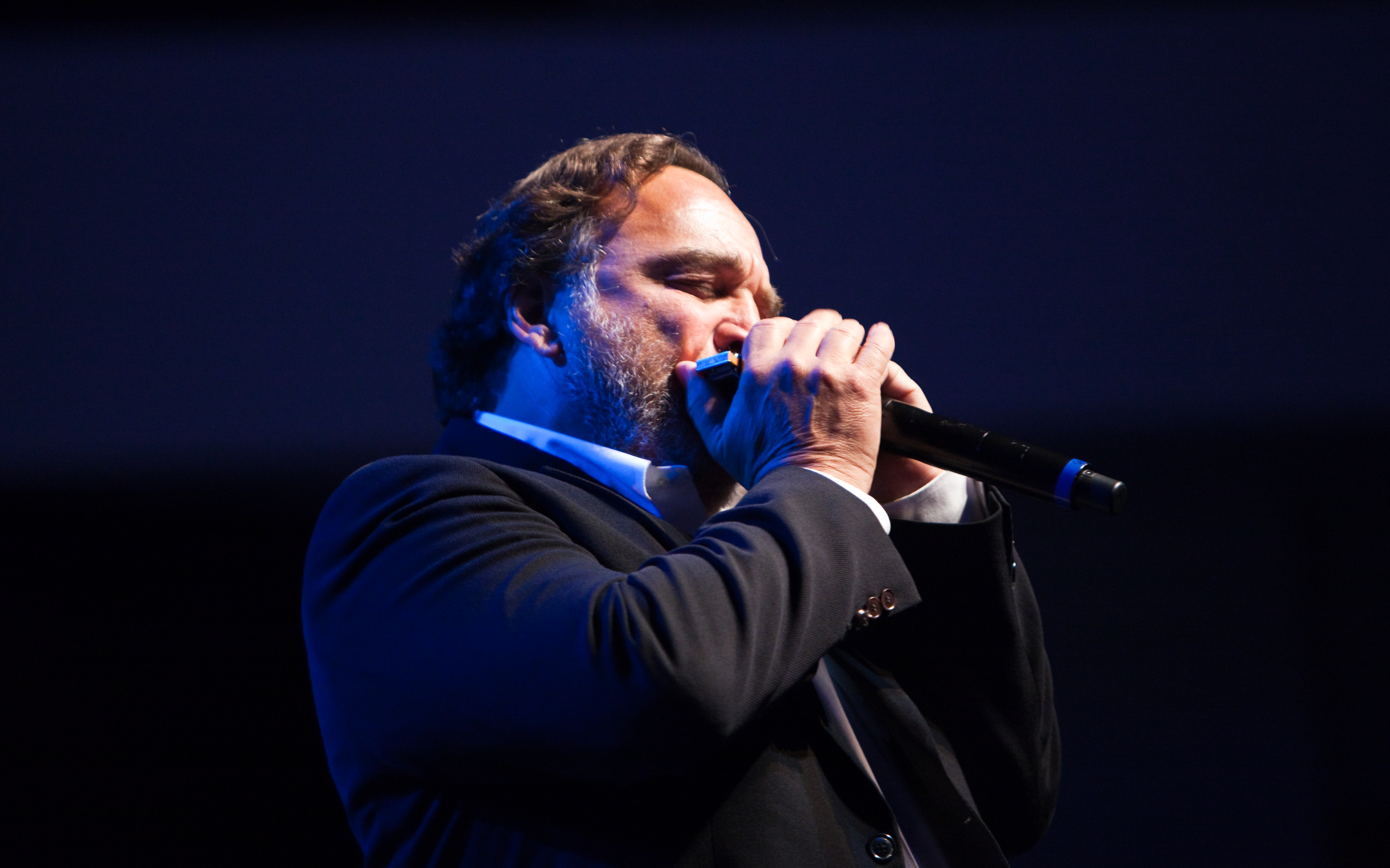 A performer in a dark suit sings into a microphone on a blue-lit stage.