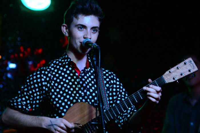 Young man in polka-dot shirt playing acoustic guitar and singing into a microphone on a dark stage.