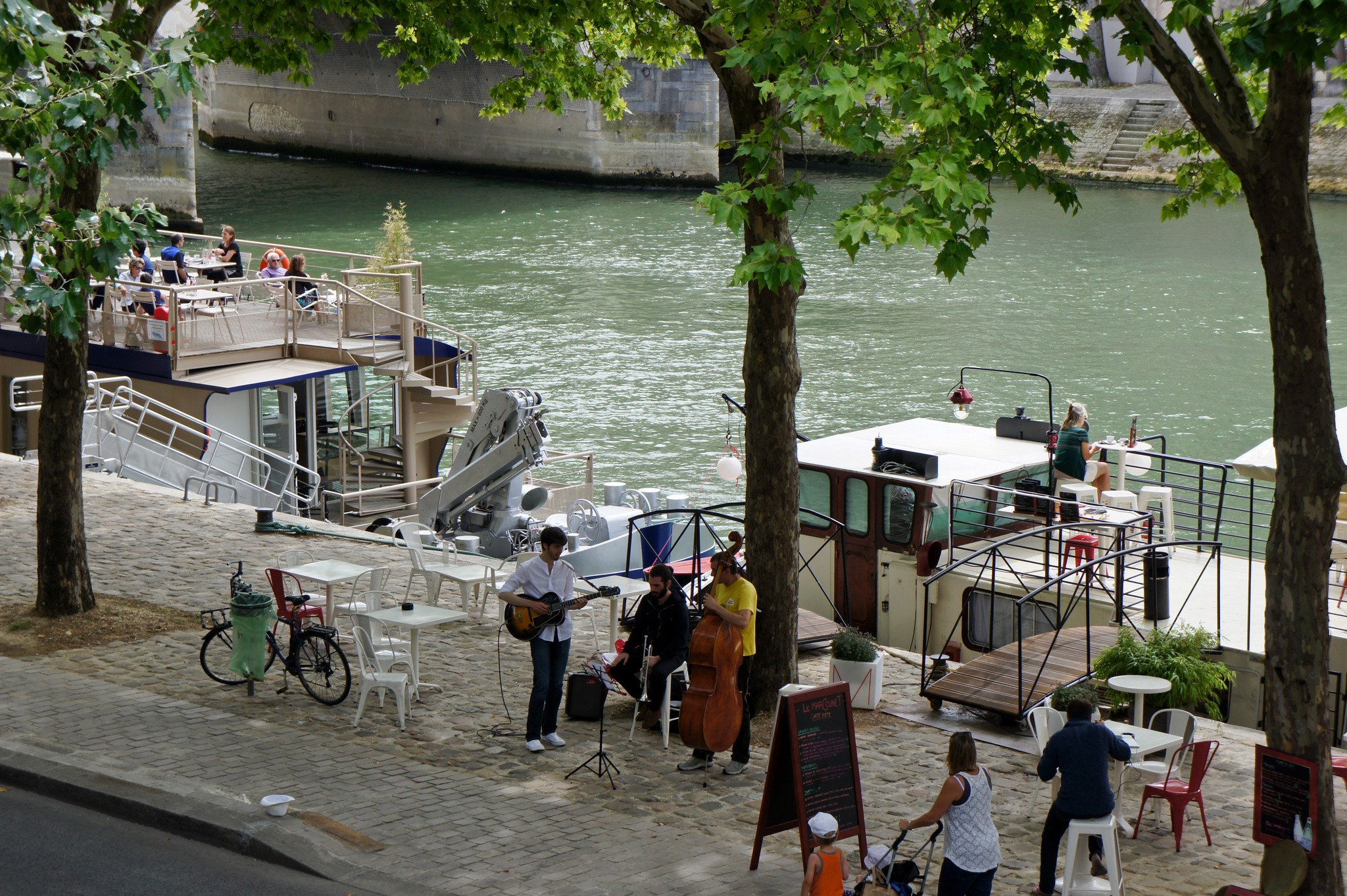 Musicians play on a cobblestone riverside quay in Paris with houseboats in the background.