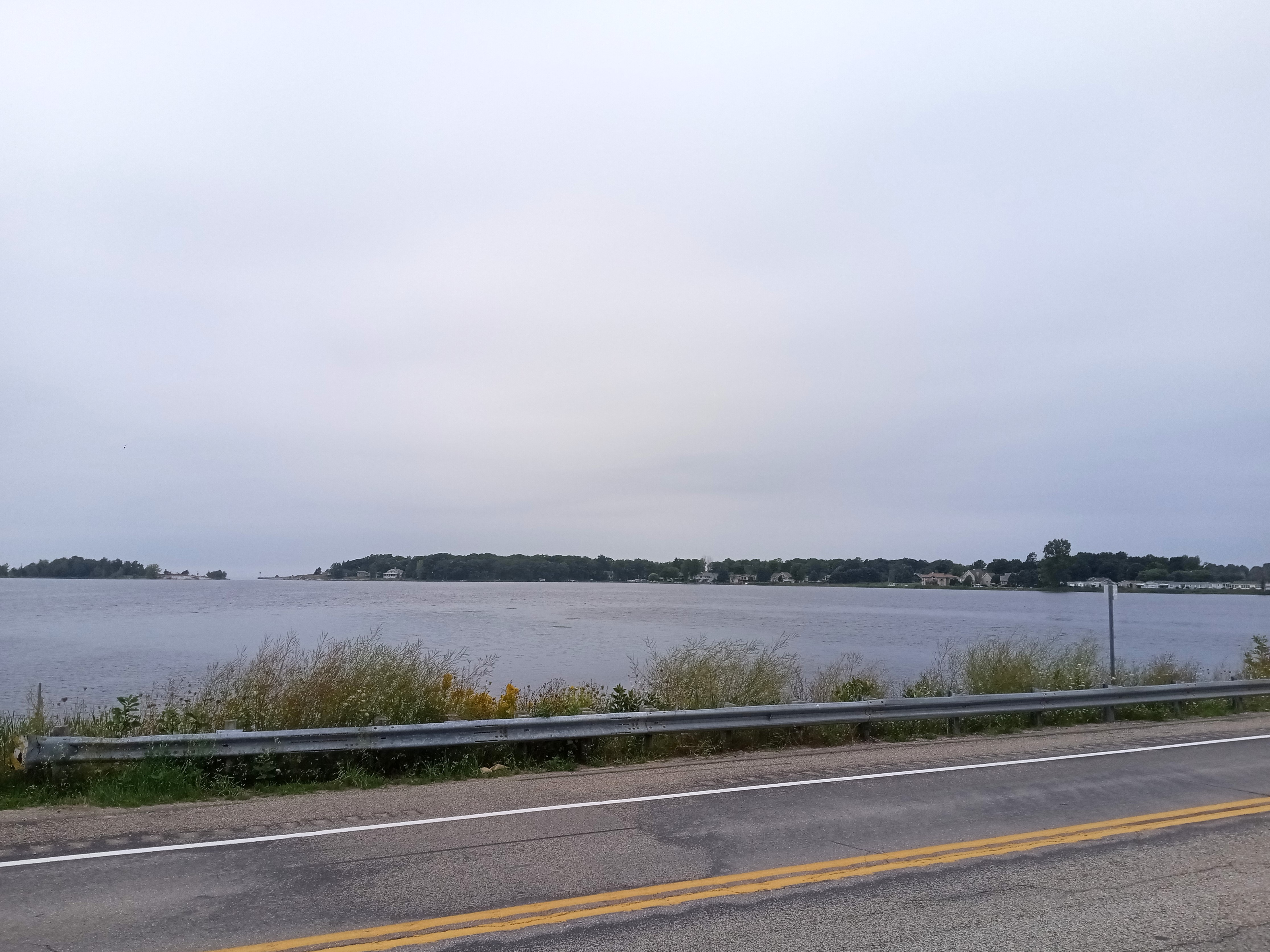 Calm lake with tree-lined far shore viewed from a paved road with a metal guardrail.