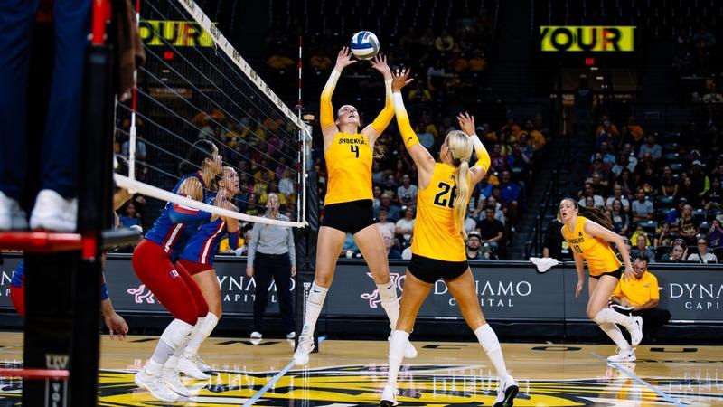 Two volleyball players in gold uniforms compete at the net against opponents in blue uniforms.