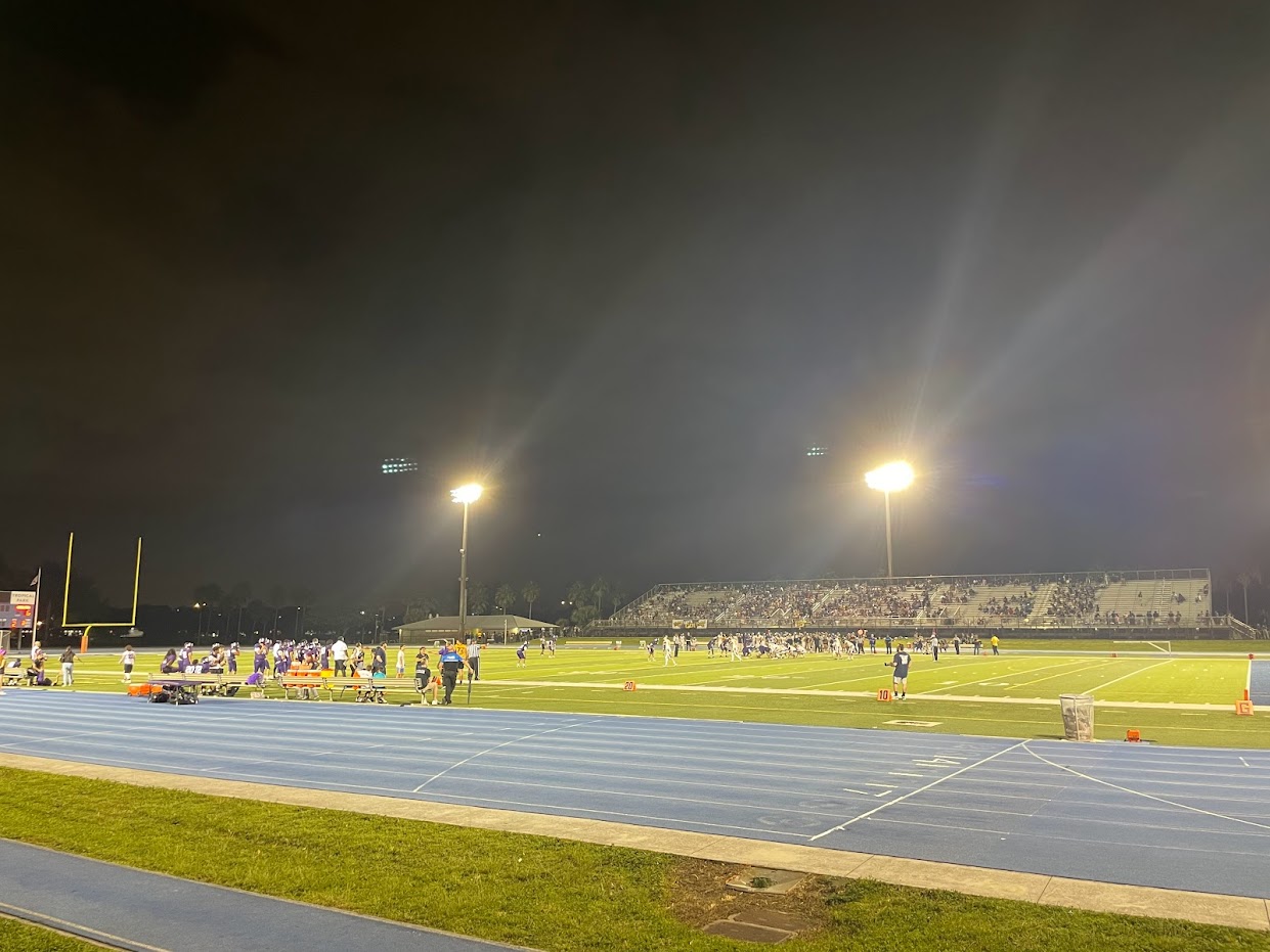 High school football game under stadium lights at night, players on field, fans in bleachers.