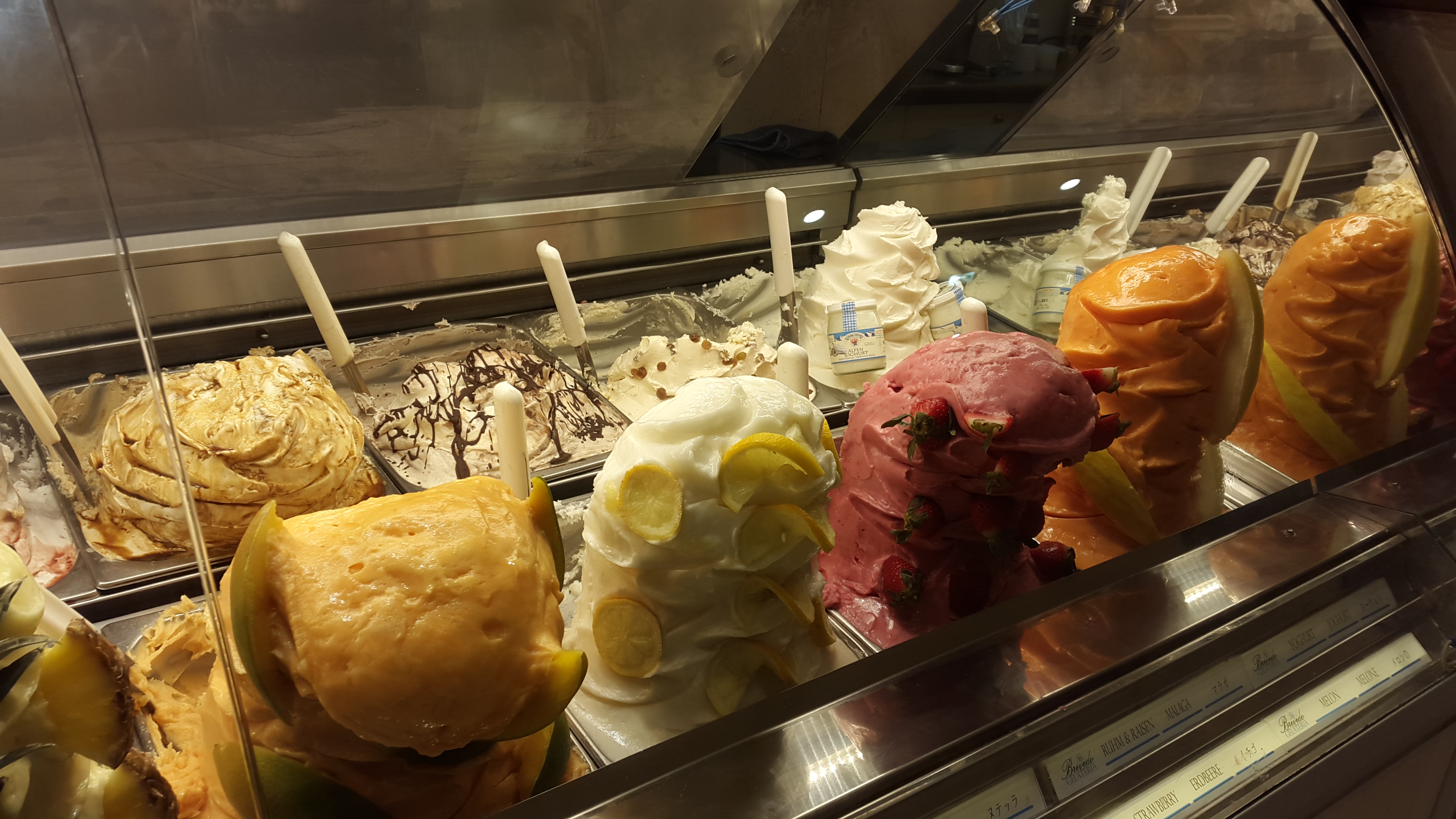 Large mounds of colorful gelato garnished with fruit slices in a glass display case.