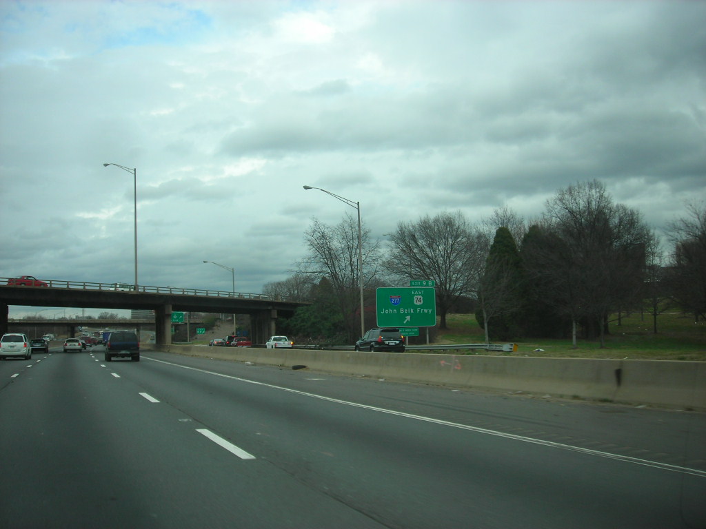 Multi-lane interstate highway with an overpass bridge and highway exit sign under an overcast sky.