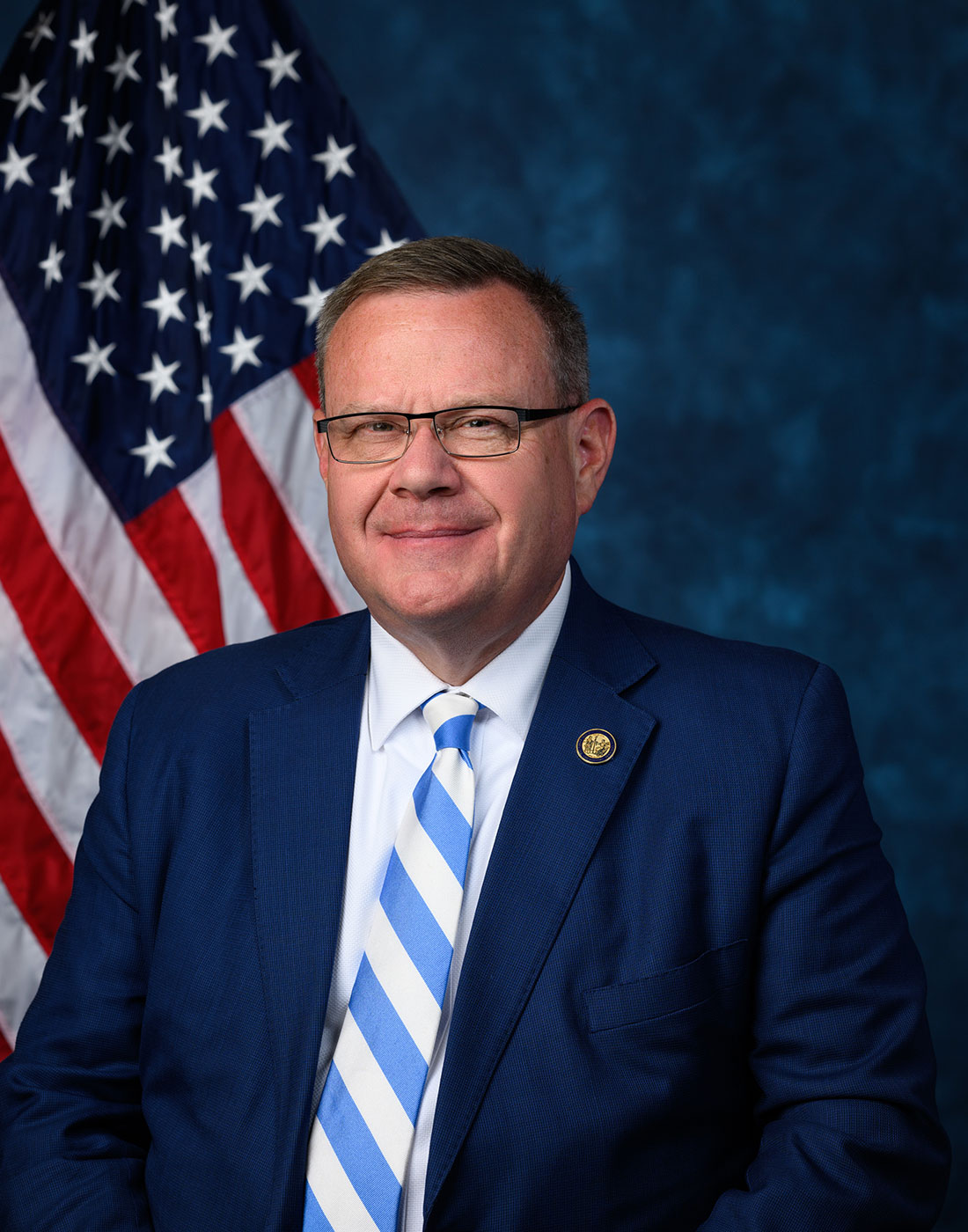 Man in navy blue suit and striped tie poses before an American flag in an official congressional portrait.
