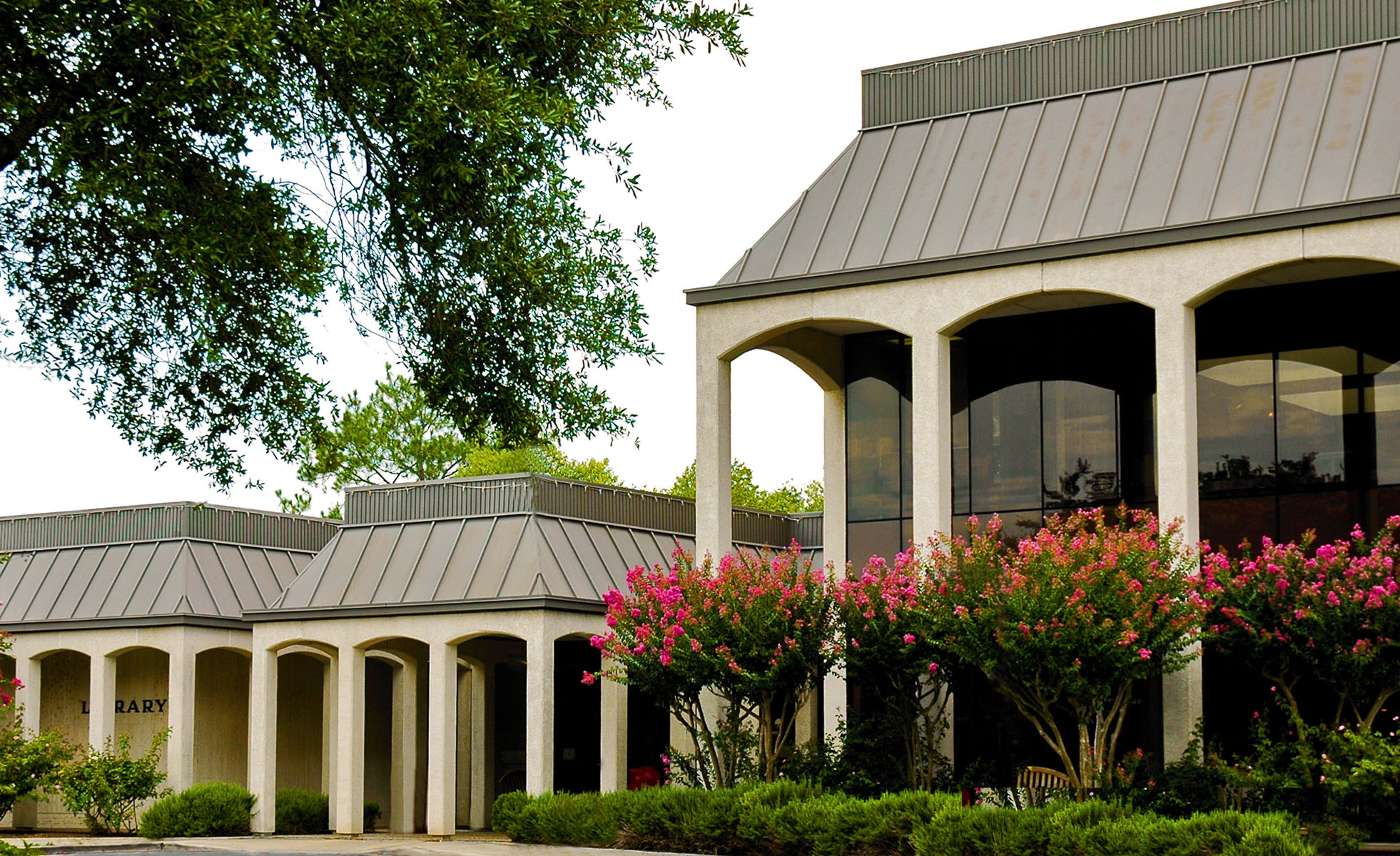 Exterior of a two-story public library with arched colonnades and blooming pink shrubs.