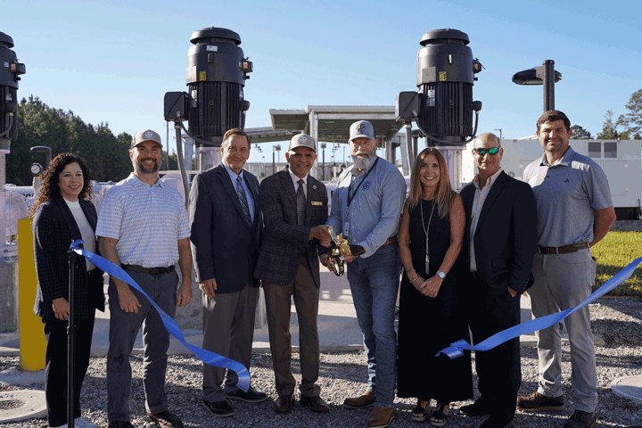 Group of eight people holding a blue ribbon in front of large industrial pump equipment outdoors.
