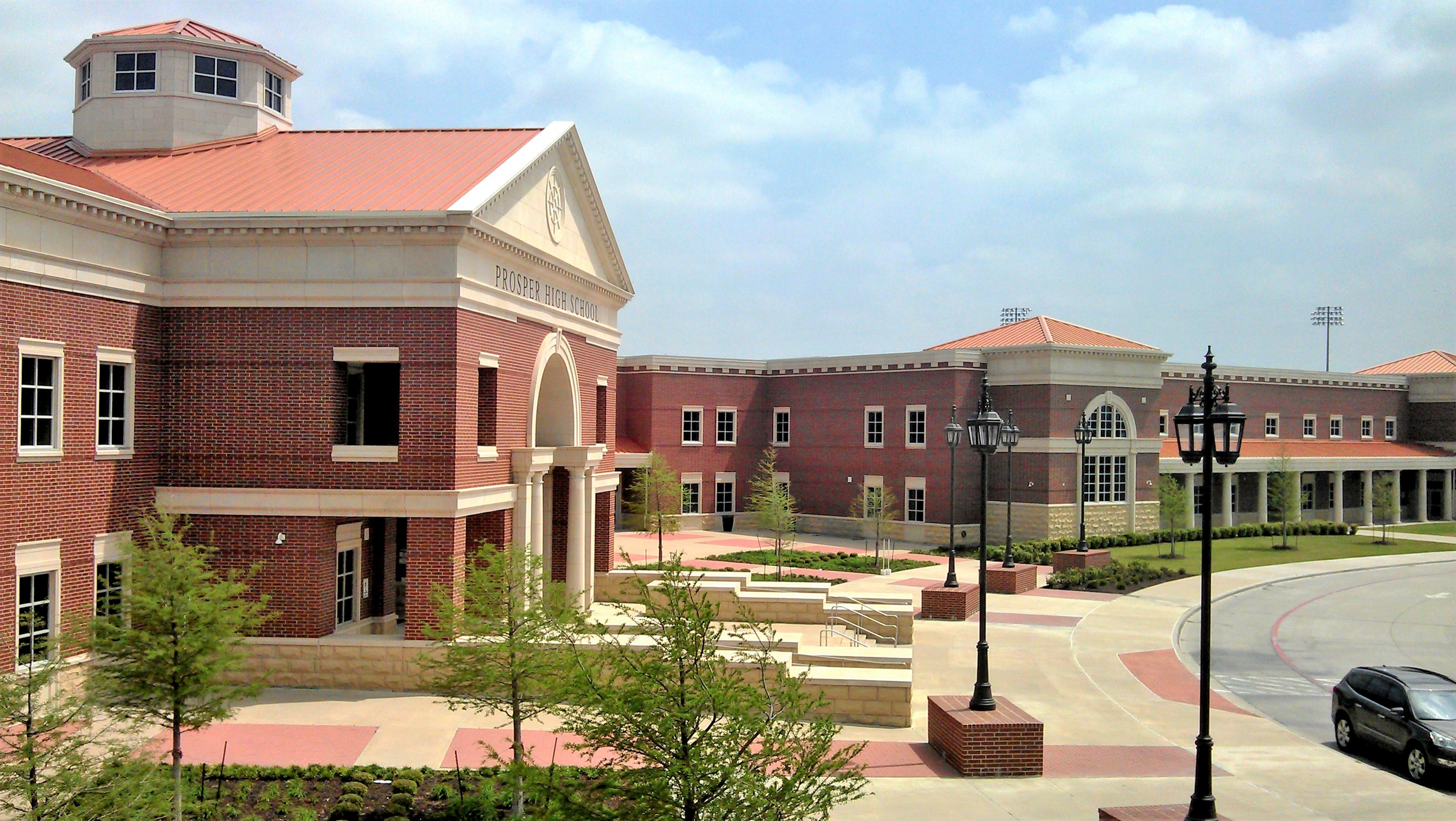 Large red-brick school building with arched entrance, lamp posts, and paved courtyard.