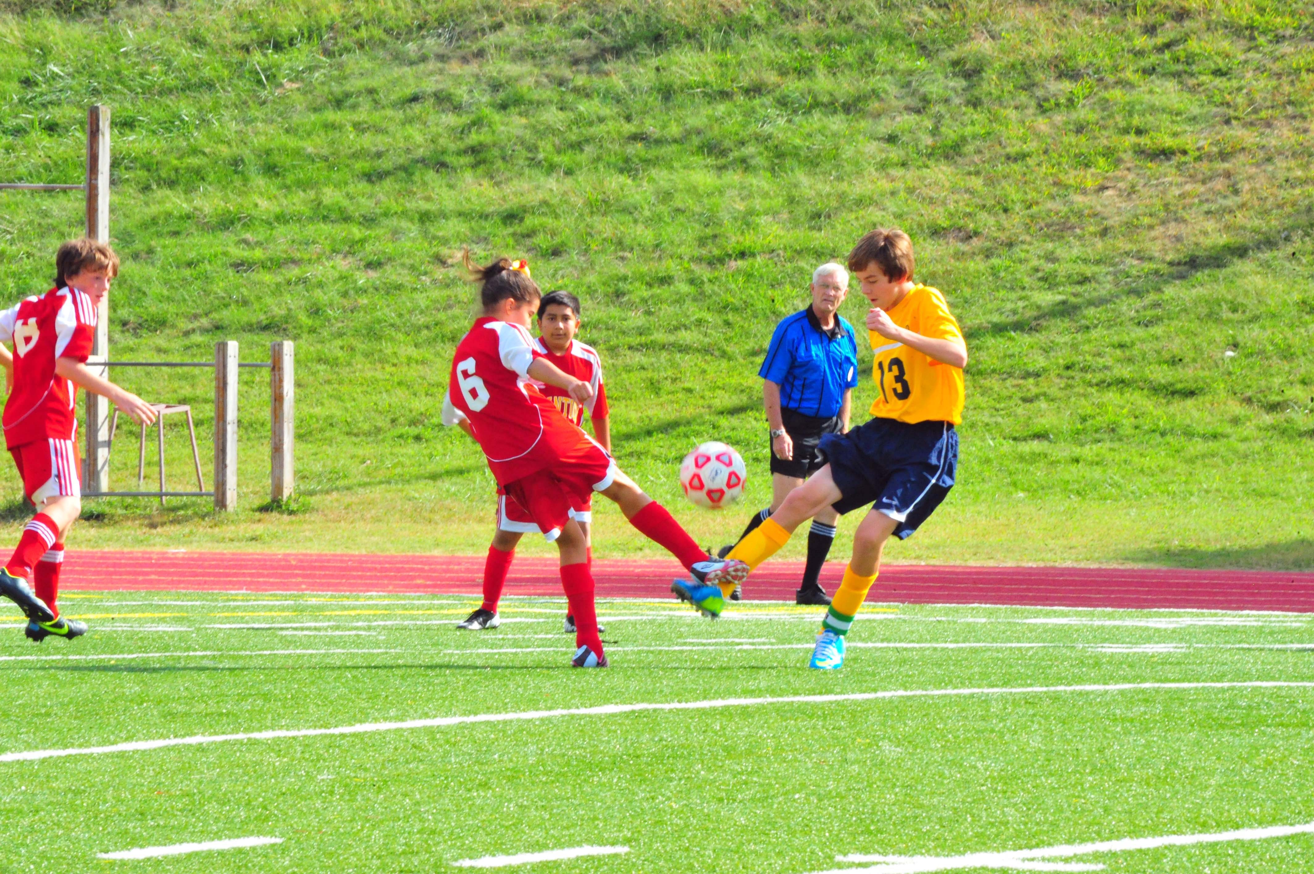 Youth soccer players in red and yellow uniforms compete for the ball on a grass field with a referee nearby.