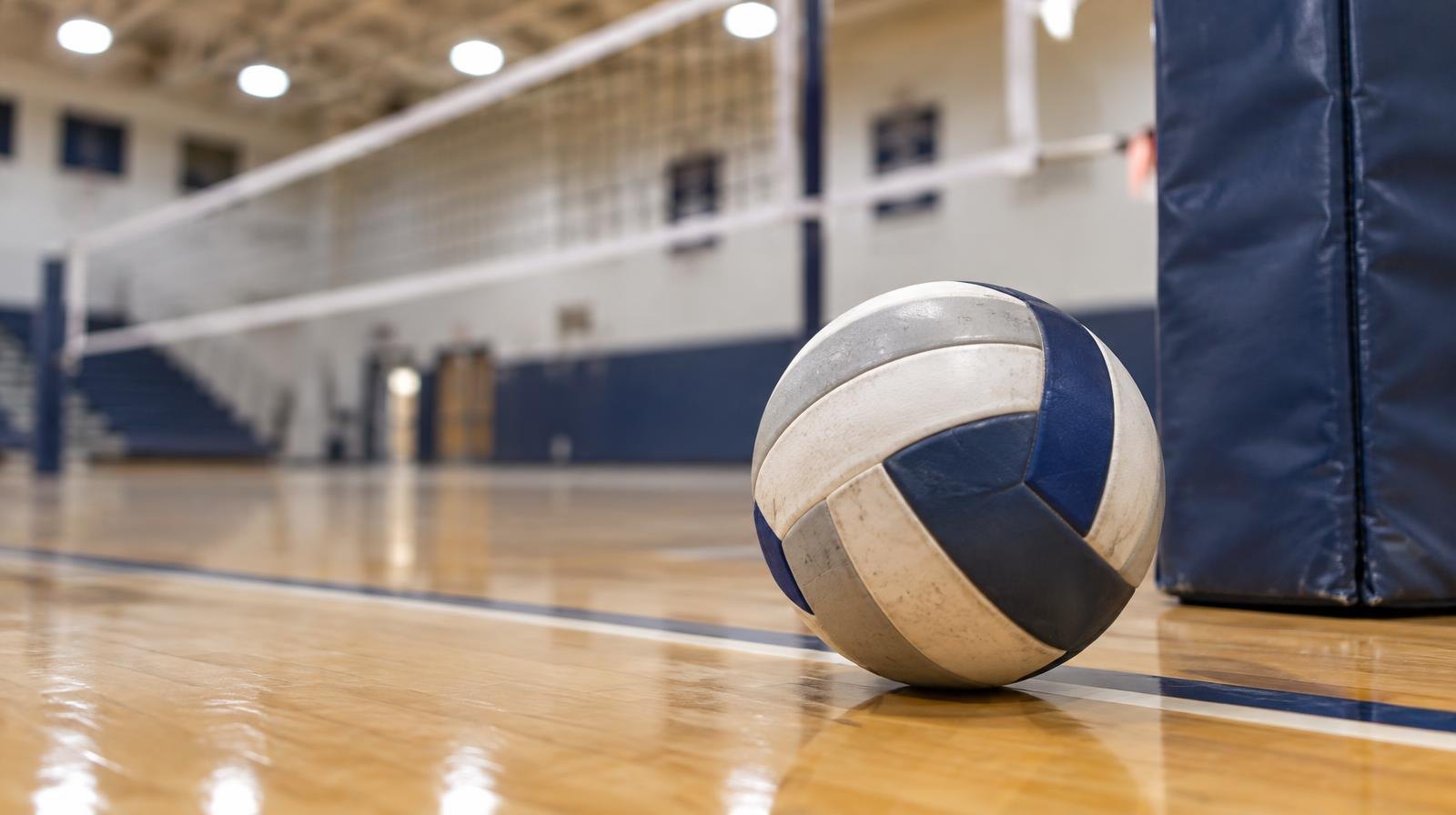 A volleyball resting on a hardwood gymnasium floor near a net post.