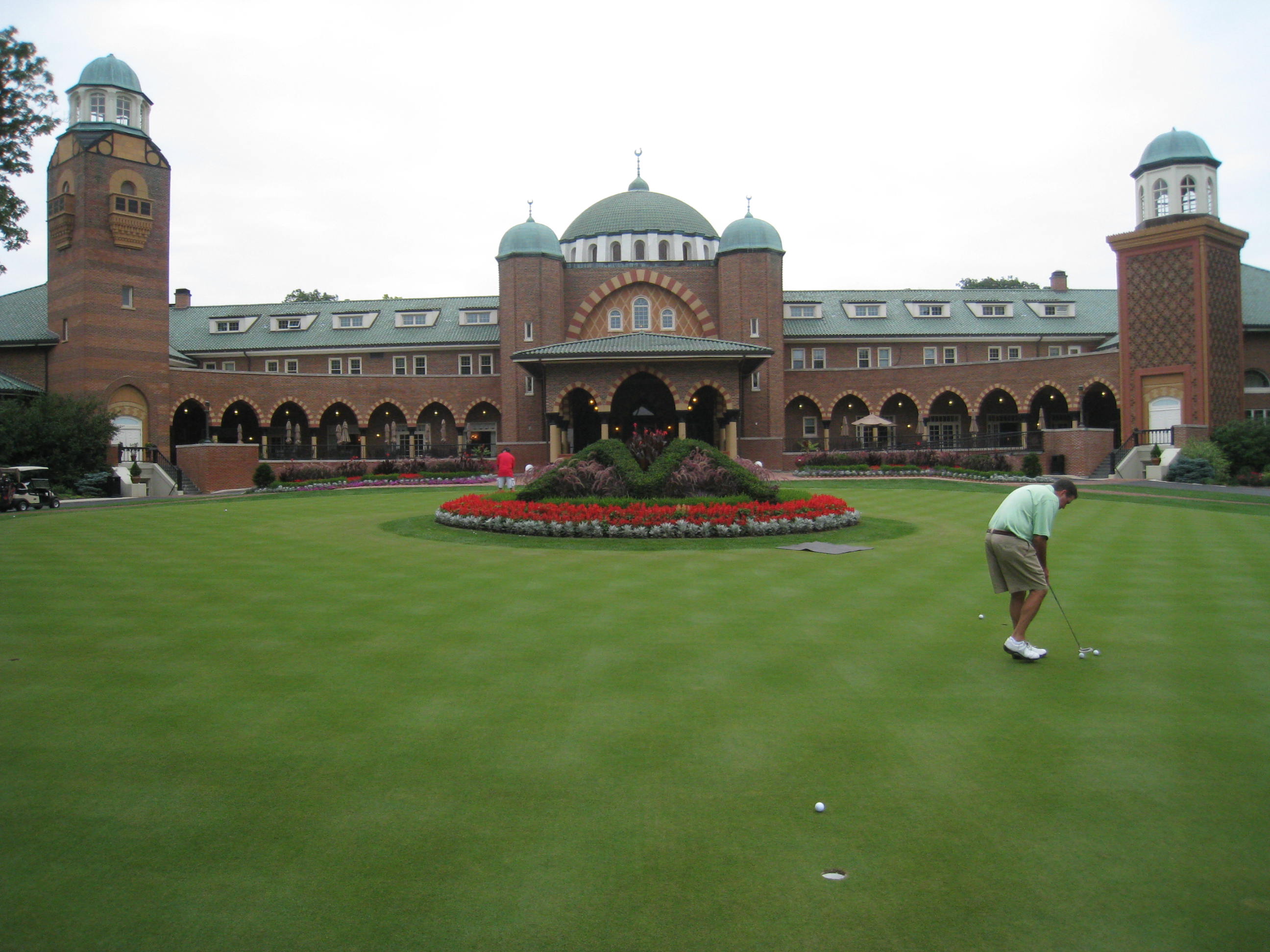 Golfer on a practice putting green in front of a brick clubhouse with green domed roofs.