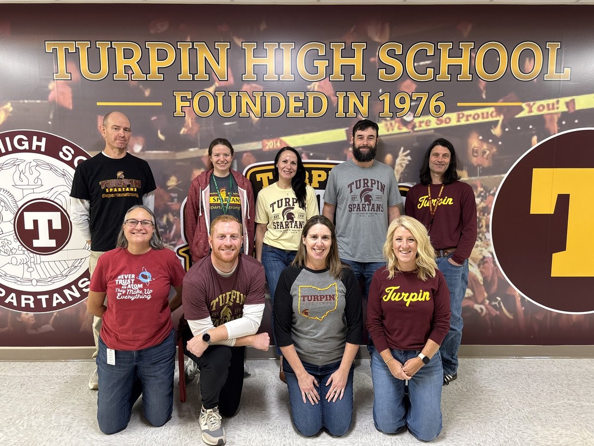 Group of nine people posing in two rows in front of a school wall display.