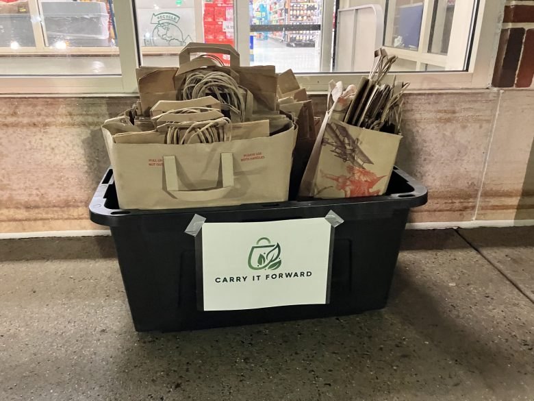 A black bin labeled Carry It Forward overflows with brown paper grocery bags.