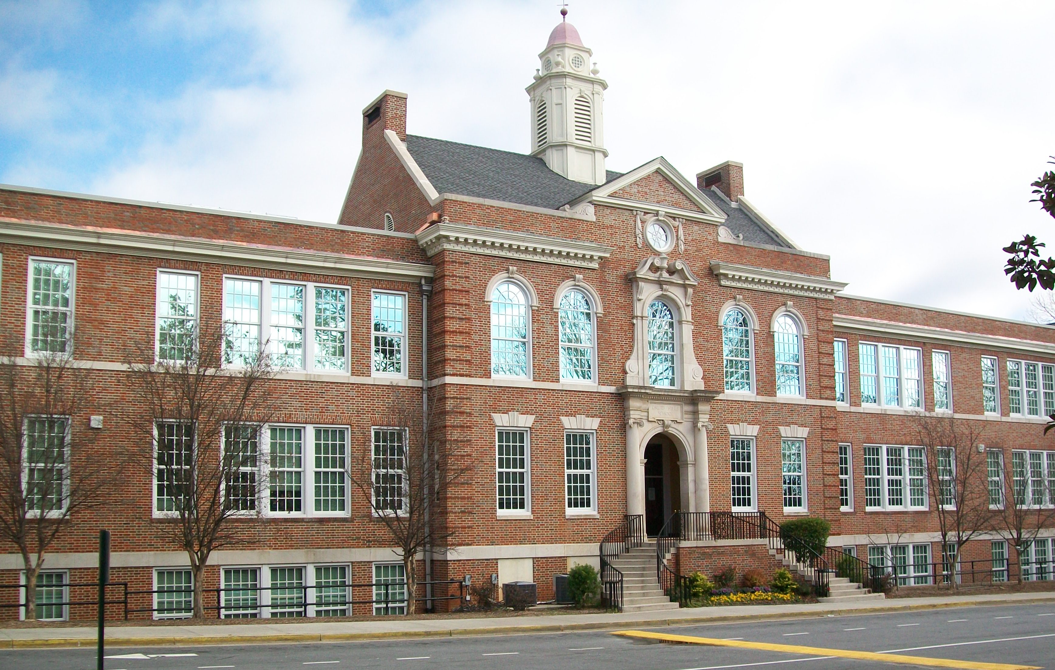 Three-story red brick school building with a white cupola and arched entrance, bare trees in front.