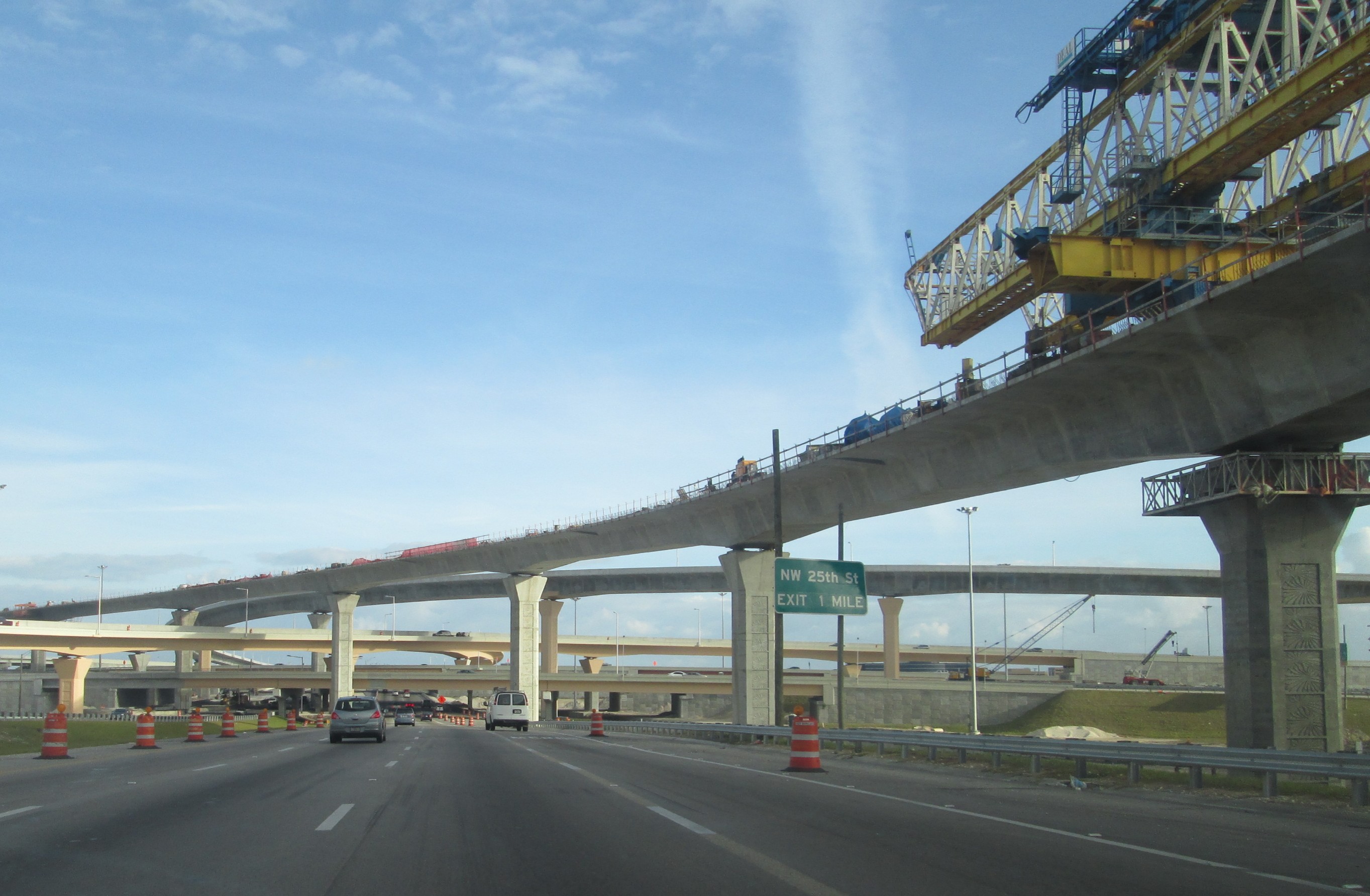 Multi-level highway interchange under construction with a yellow crane and orange traffic barrels.