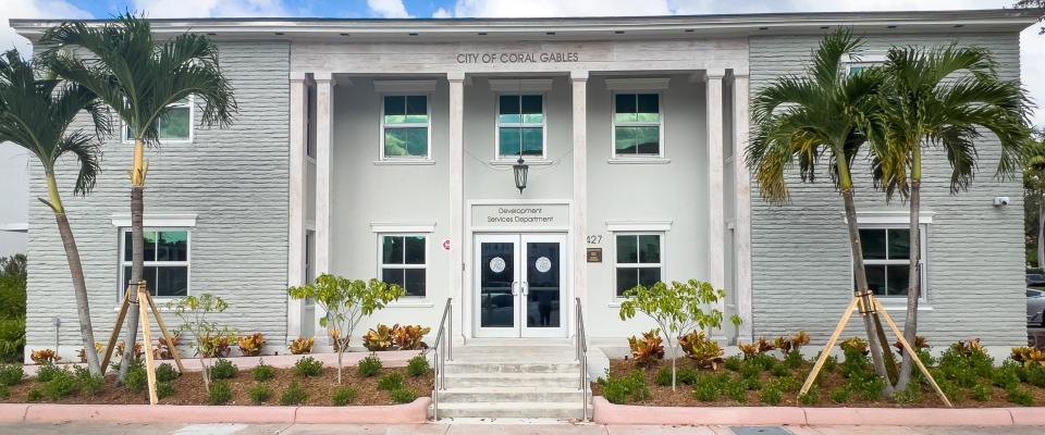 White two-story government building with columns and palm trees under a blue sky.