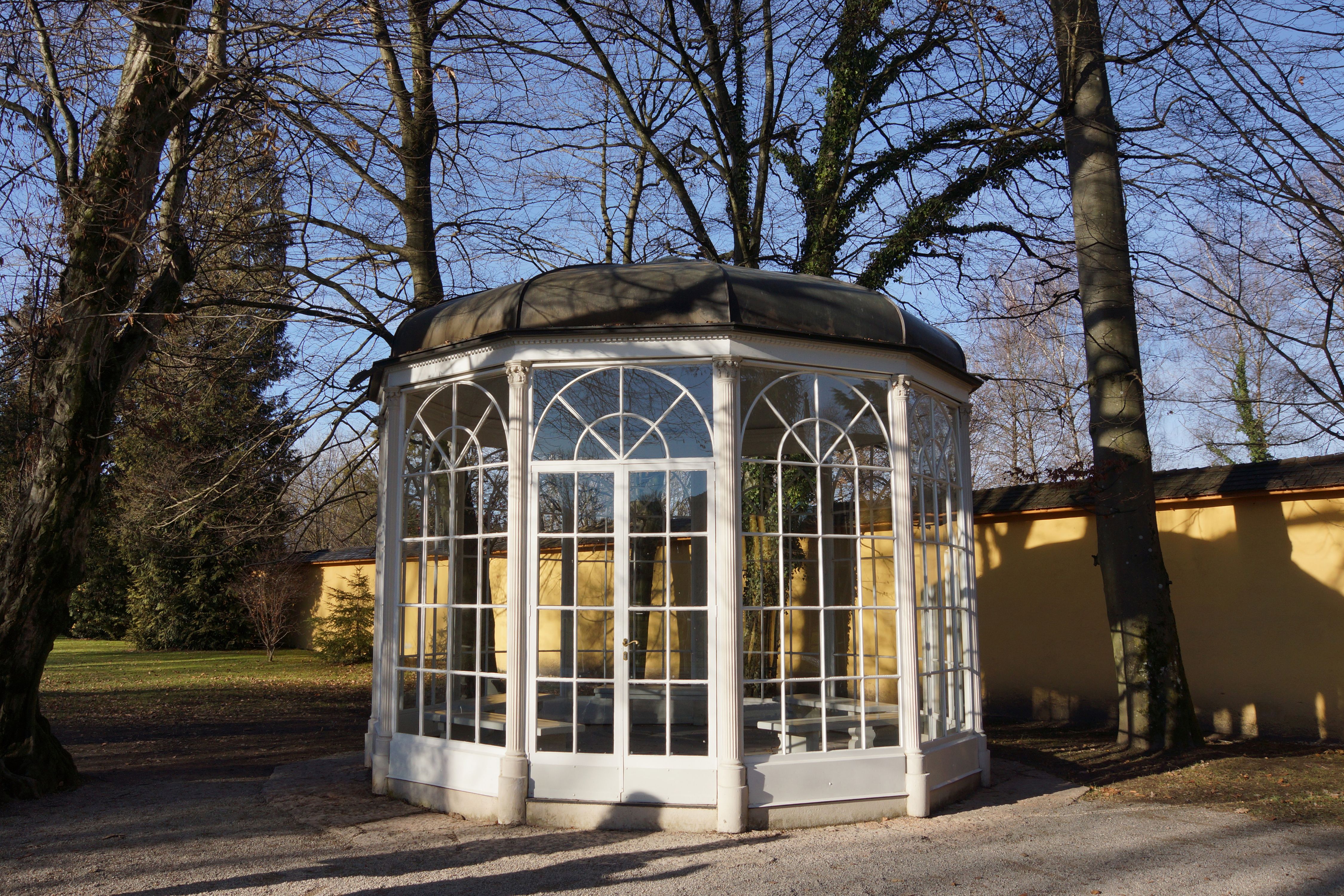 An octagonal white-framed glass gazebo surrounded by bare autumn trees in a park.