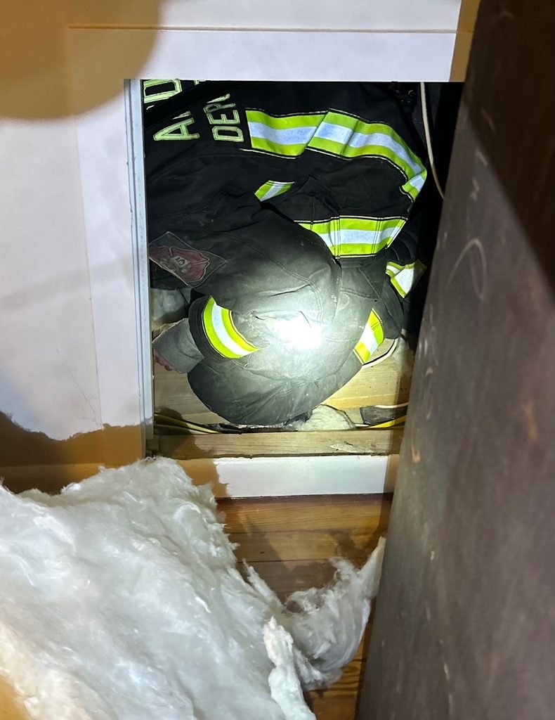 Firefighter in turnout gear crouched inside a narrow crawlspace with insulation visible.
