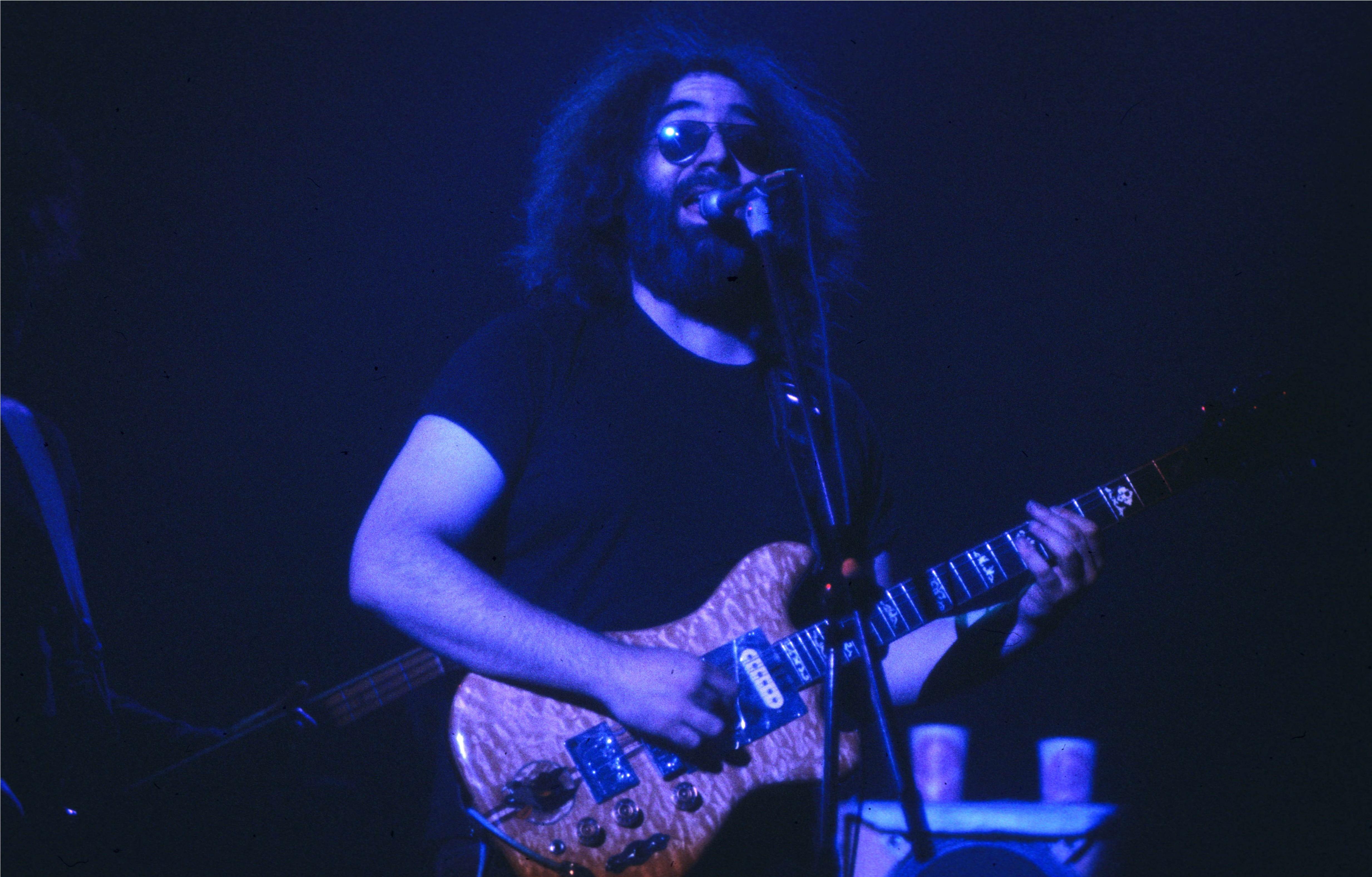 Guitarist performing on stage under blue lighting, playing an ornate electric guitar at a microphone.