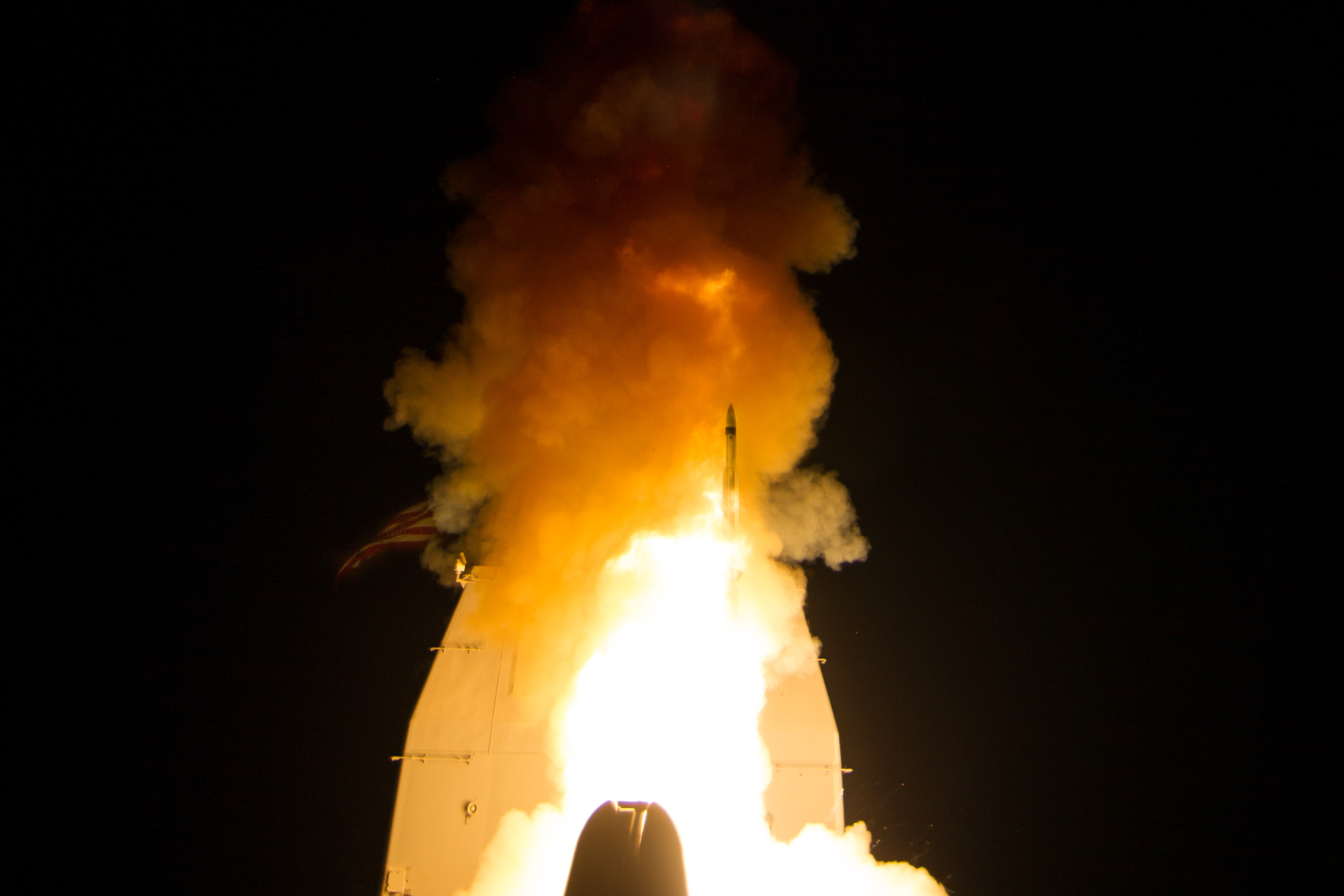 A missile launches from a Navy warship with a large plume of fire and smoke at night.