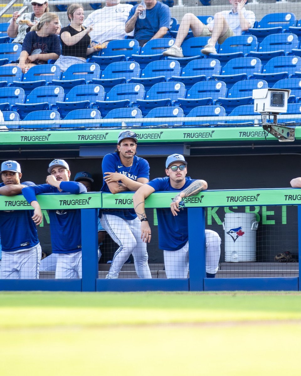 Several baseball players in blue uniforms lean on a dugout railing in a minor league stadium.