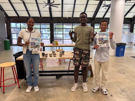 Three young people standing indoors holding printed materials at a workshop table.