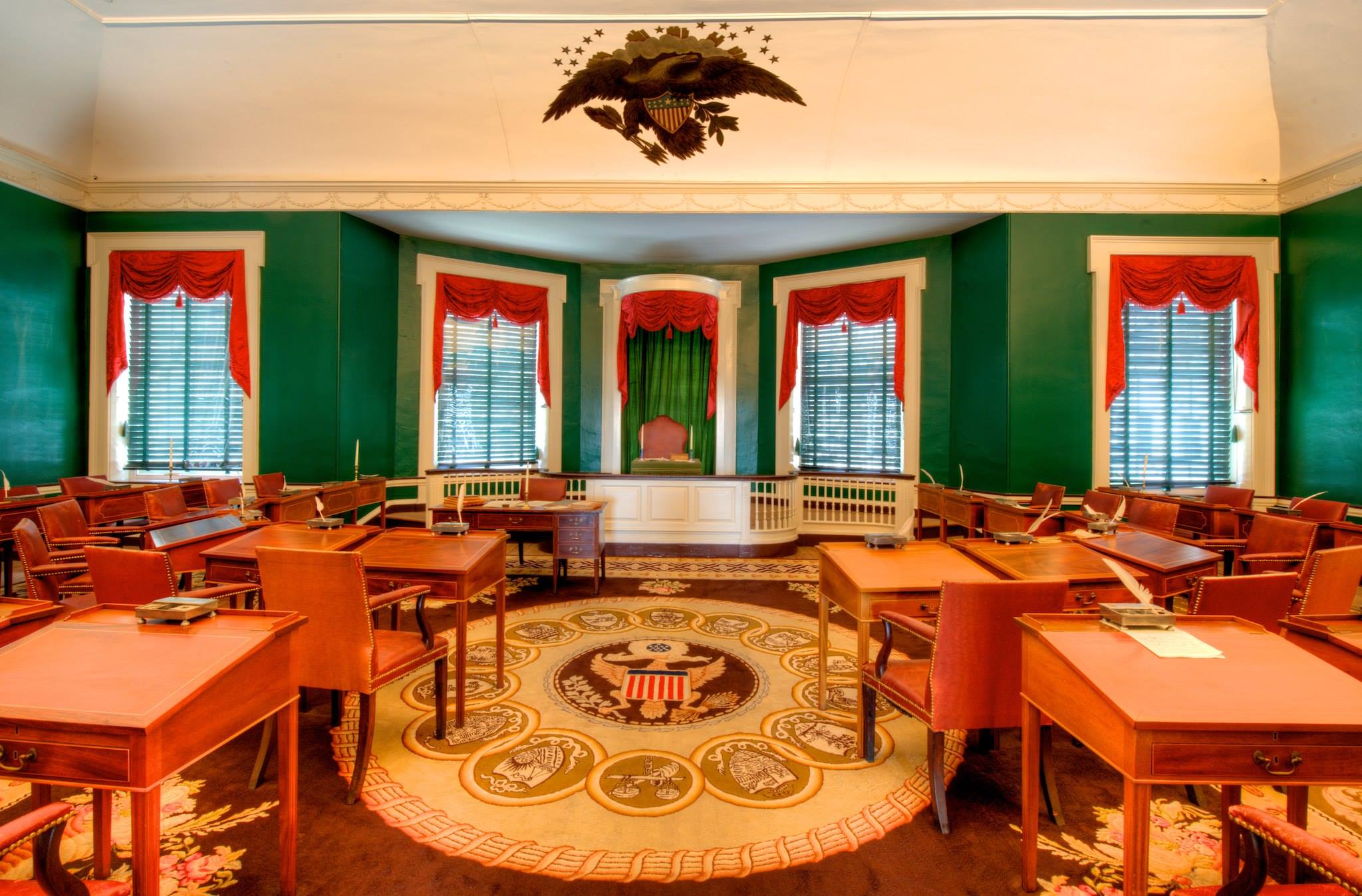 Historic legislative chamber with semicircular wooden desks, eagle-seal rug, and red-draped windows.