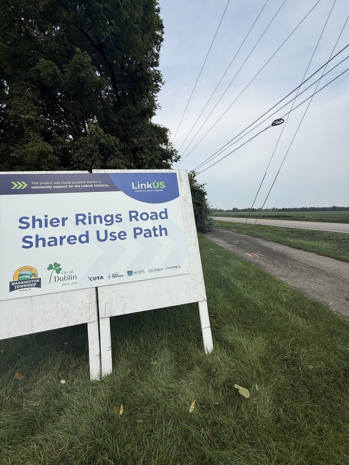 White project sign on two posts reading 'Shier Rings Road Shared Use Path' with logos, beside a grassy roadside.