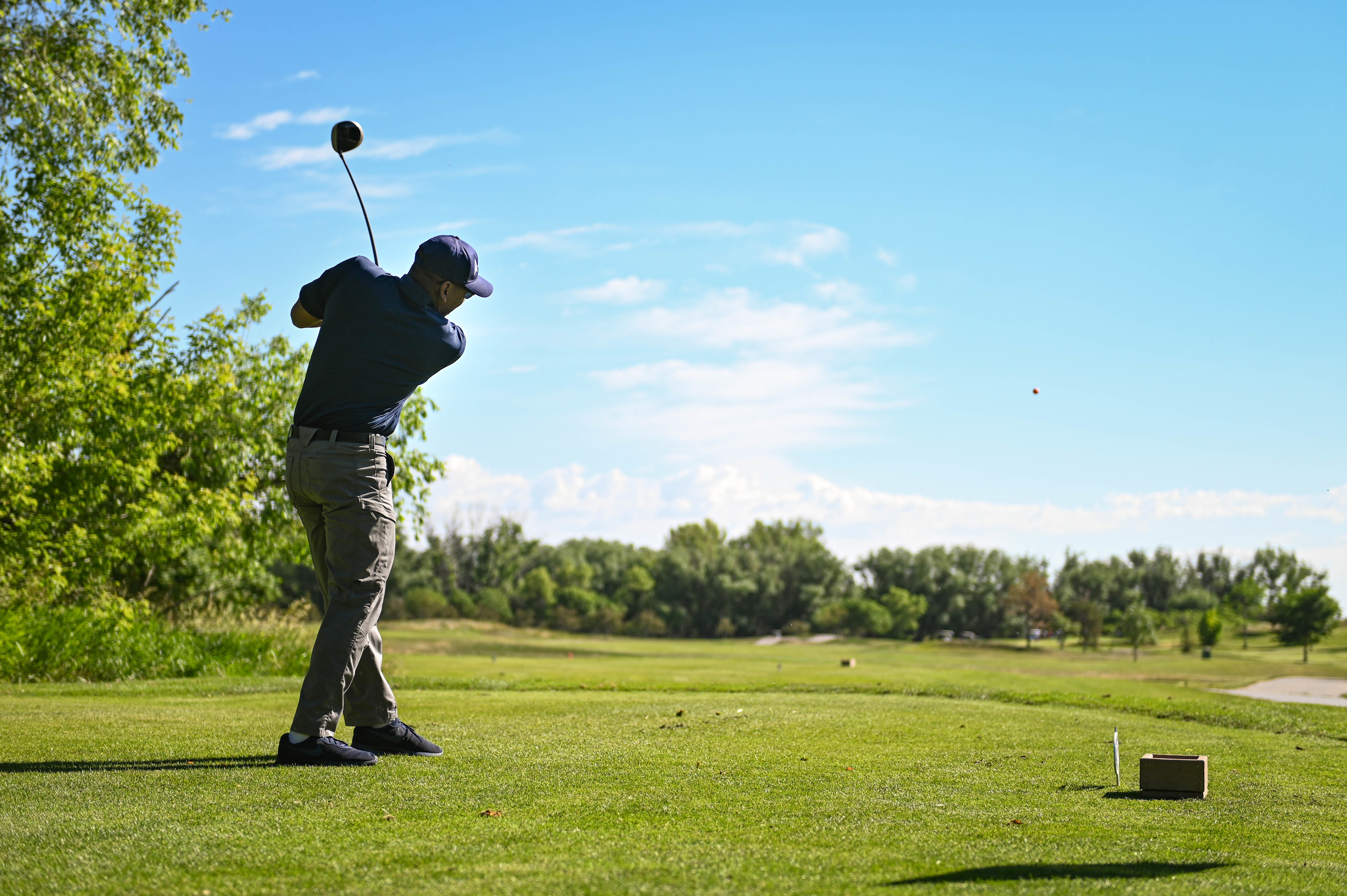 A golfer completes a full swing on a tee box, golf ball visible in the air.