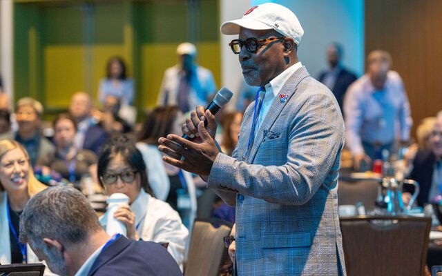 A man in a light blue blazer speaks into a microphone at an indoor forum with a seated audience behind him.