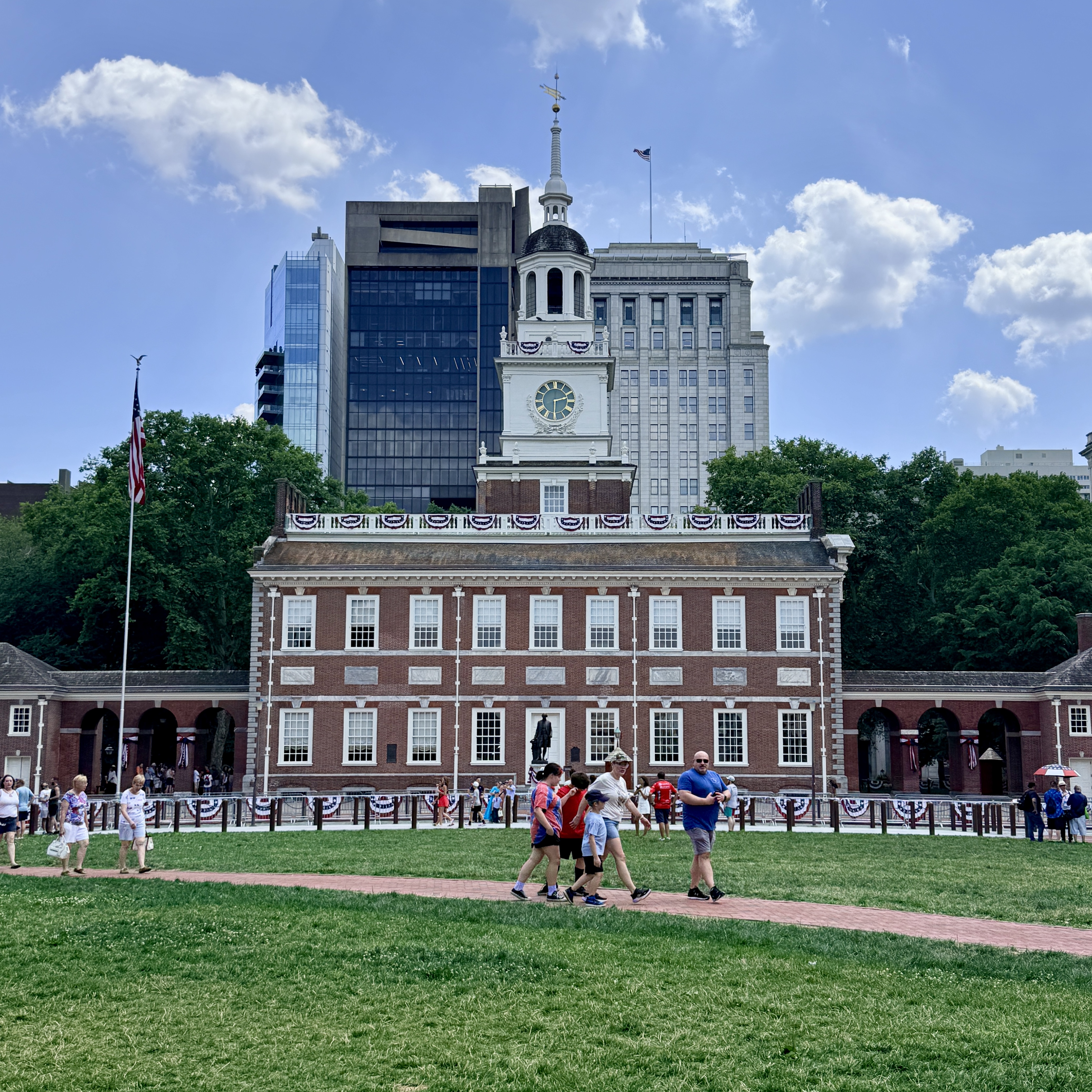 Historic red-brick building with white steeple, patriotic bunting, and a crowd on the lawn.