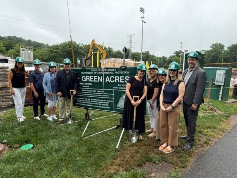 Group of officials in green hard hats holding golden shovels at a park groundbreaking ceremony.