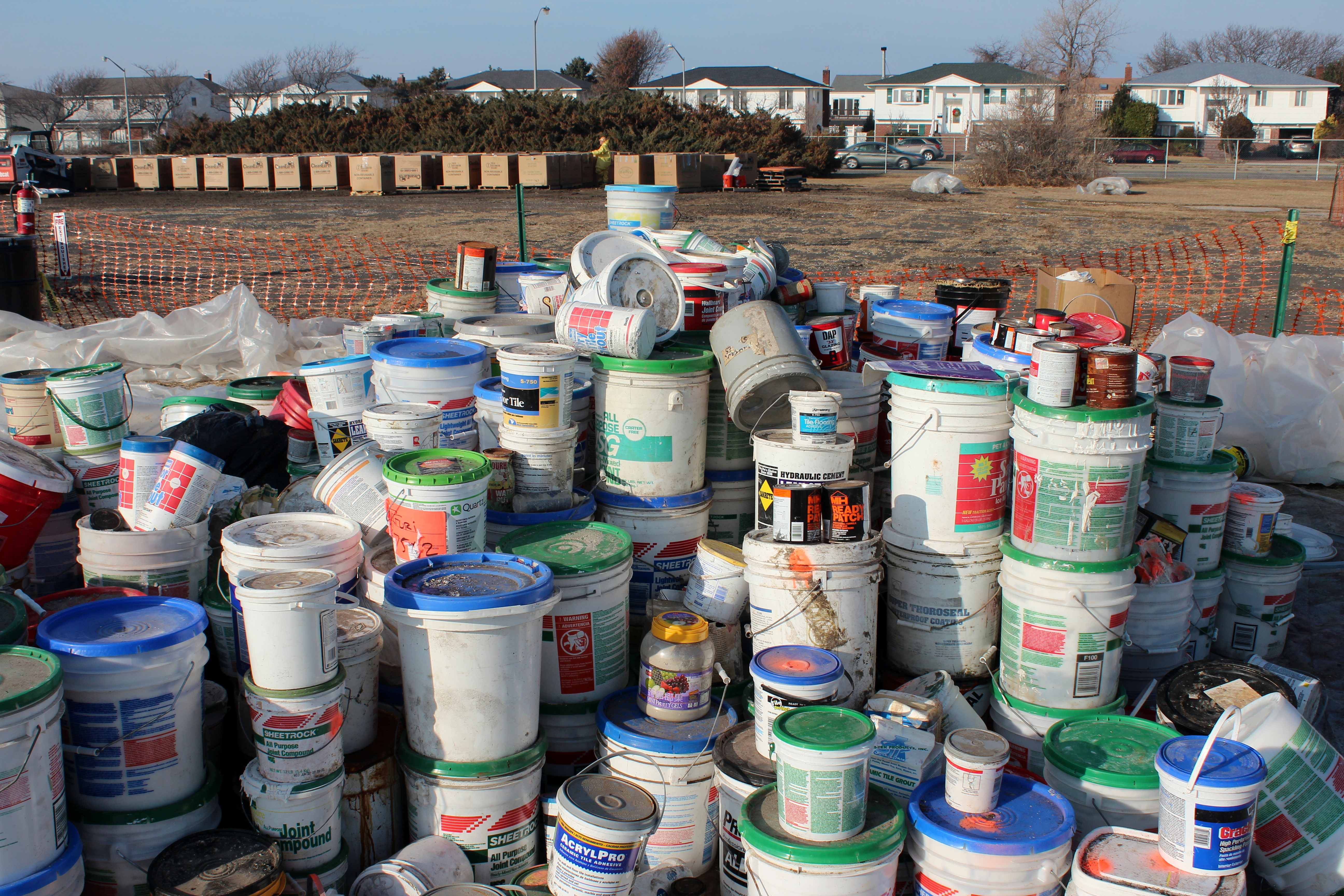 Dozens of plastic paint buckets and chemical containers stacked outdoors at a collection site.