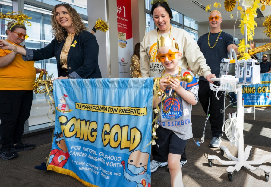 A young boy wearing gold star sunglasses and a gold crown walks past a "Going Gold" banner for Texas Children's Cancer and Hematology Center, accompanied by hospital staff in gold attire and an IV pole, during a Childhood Cancer Awareness Month event.