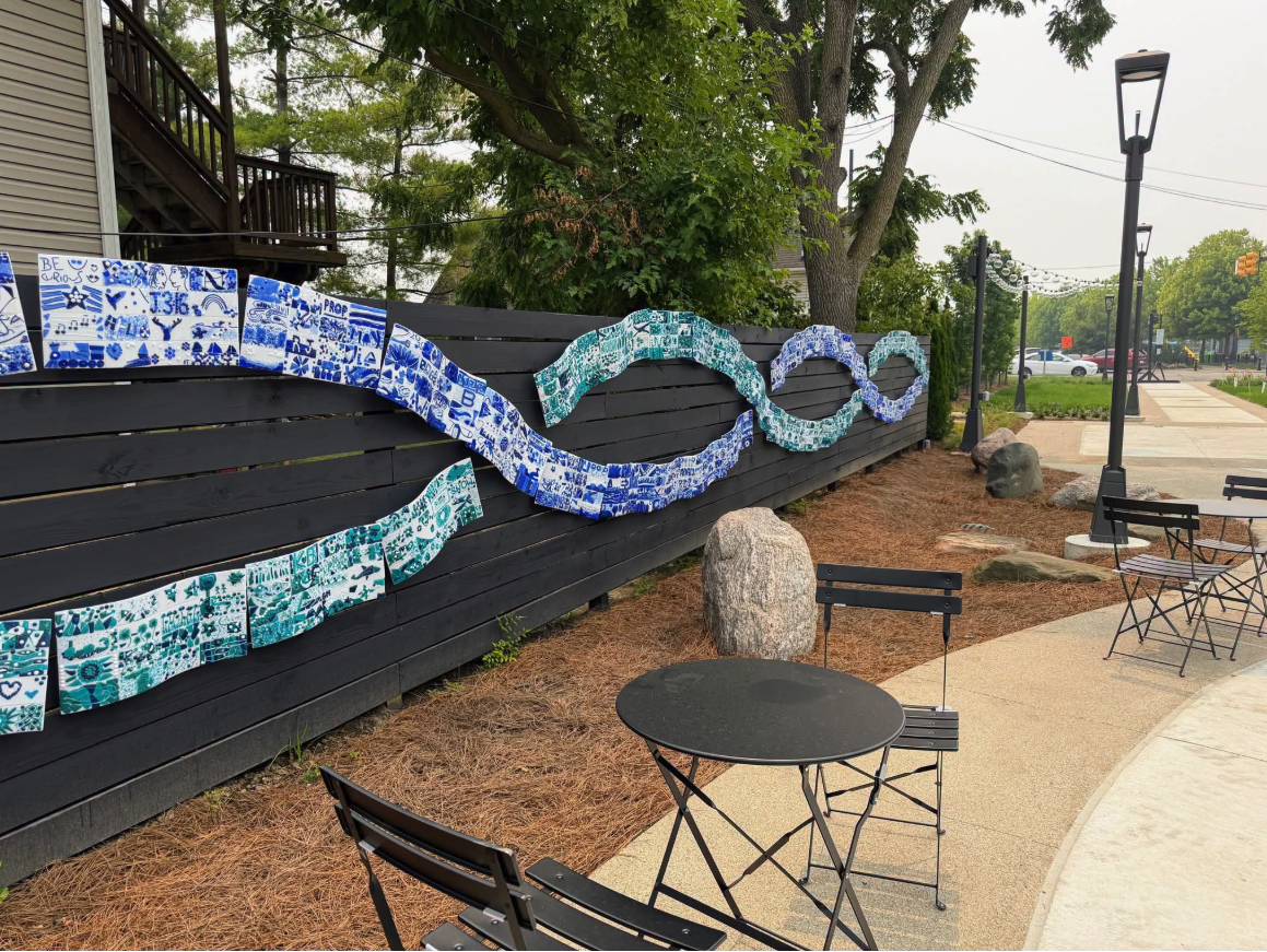The wave-shaped Make-a-Wave glass mosaic mural mounted on a black fence along the Art Park Promenade in downtown Farmington, with seating and additional community-made tiles nearby.