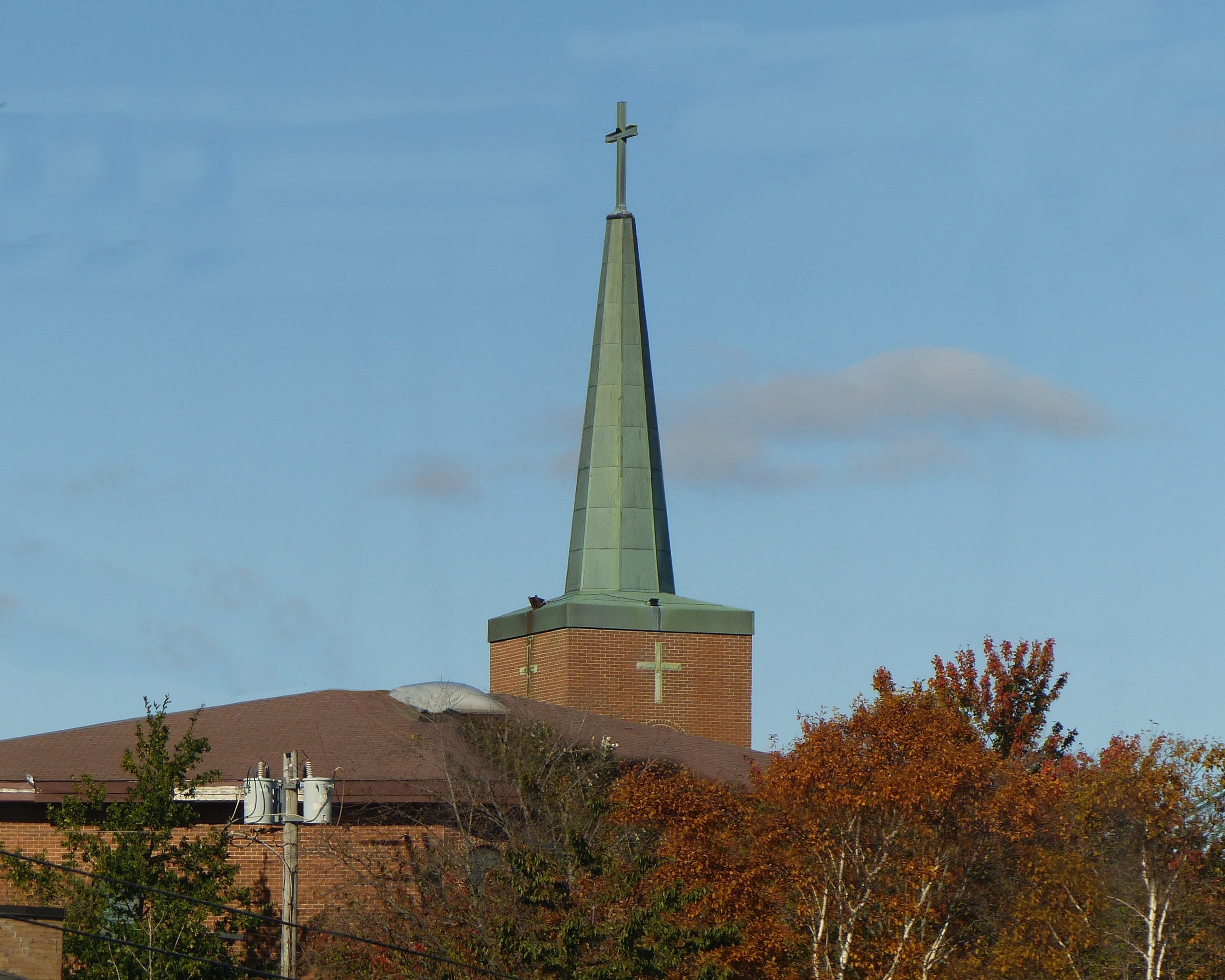 Red brick church with a green copper steeple and cross, surrounded by autumn foliage.