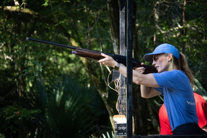 A woman in a blue cap aims an over-under shotgun at an outdoor wooded shooting range.