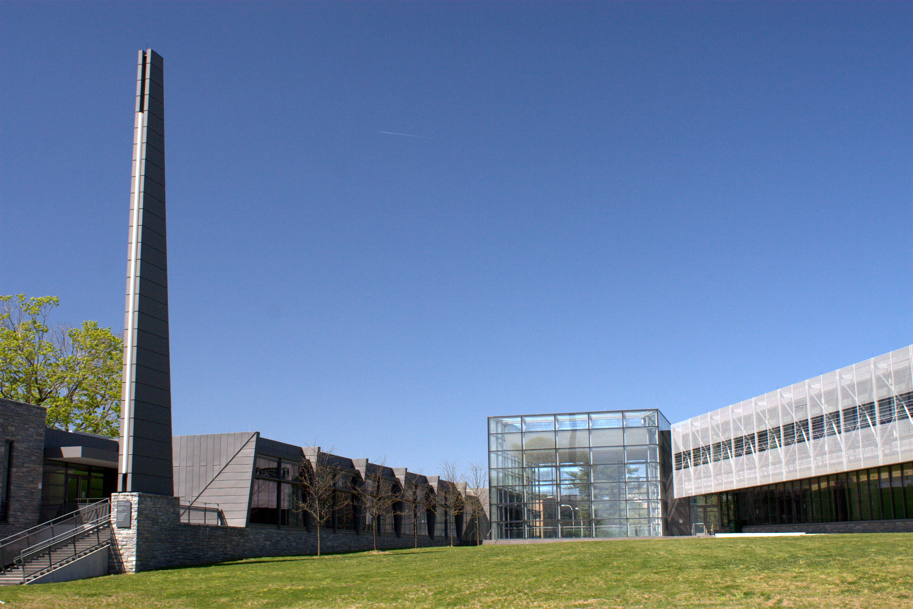 Modern glass-walled campus building with a tall tower structure on a green lawn under blue sky.