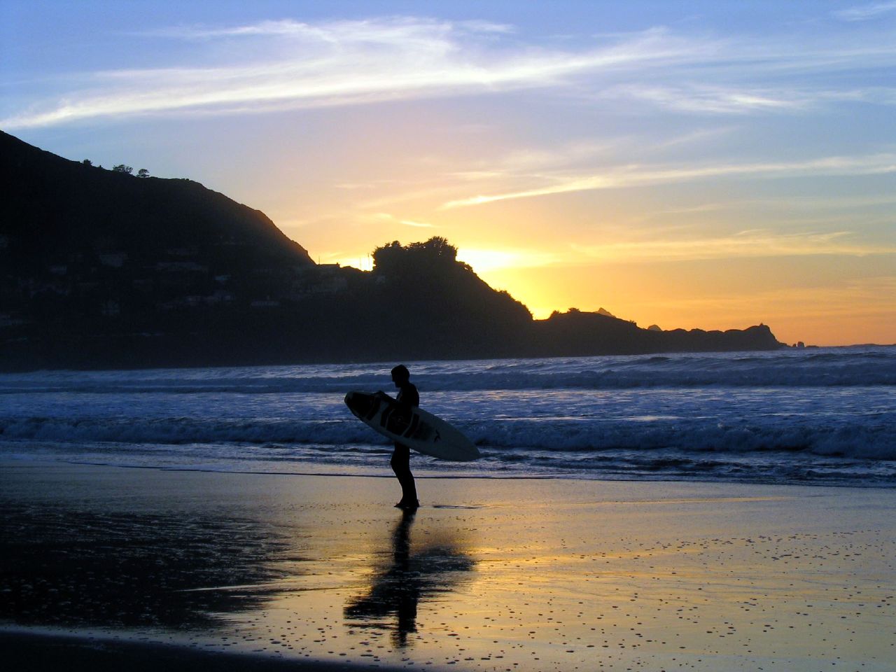 Silhouetted surfer carrying a surfboard along a beach at sunset with waves and mountains behind.