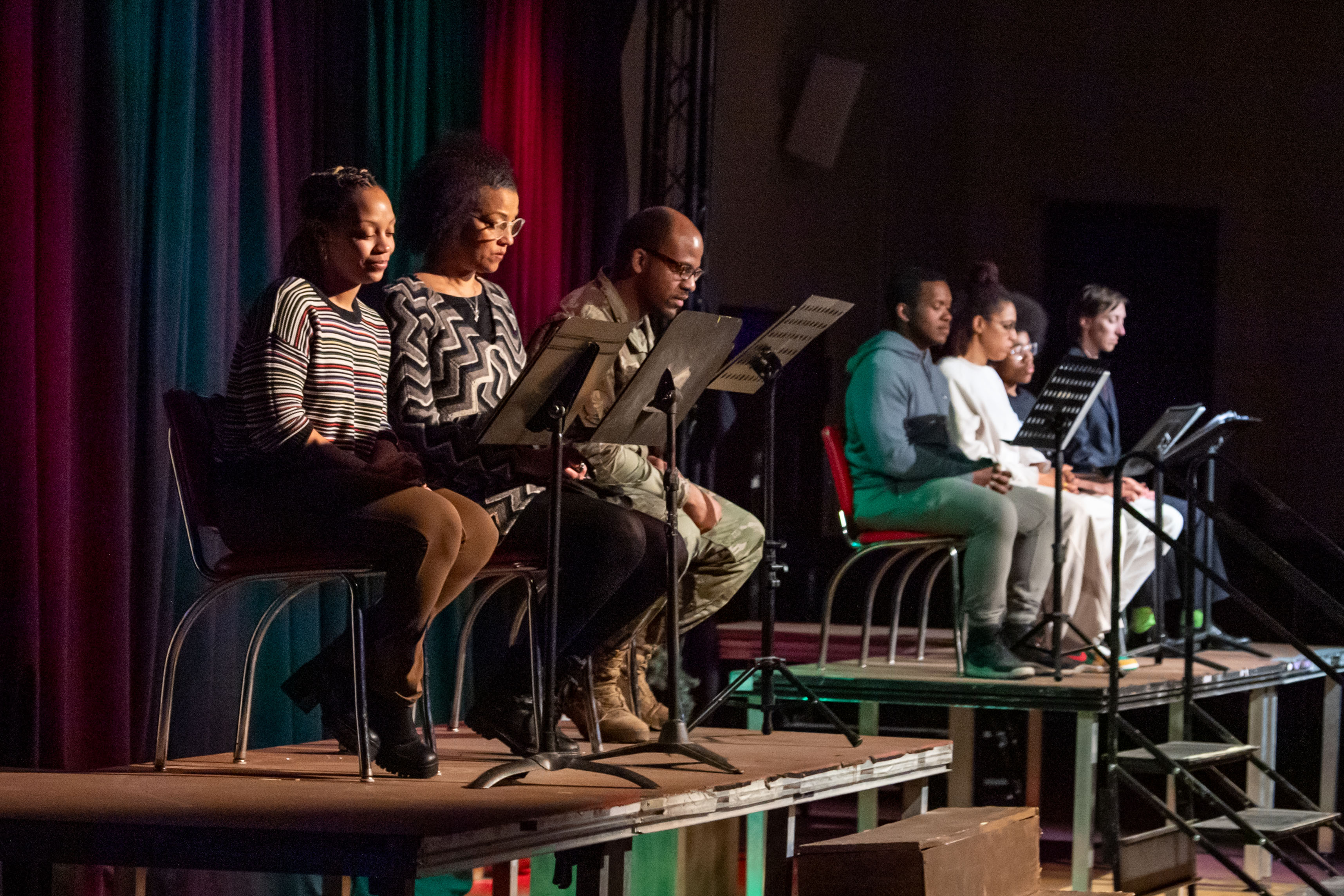 Several performers seated on stage reading scripts from music stands under colorful lighting.