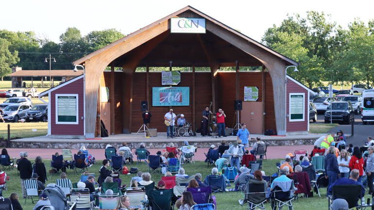 Outdoor amphitheater stage with performers and a large lawn audience at dusk.